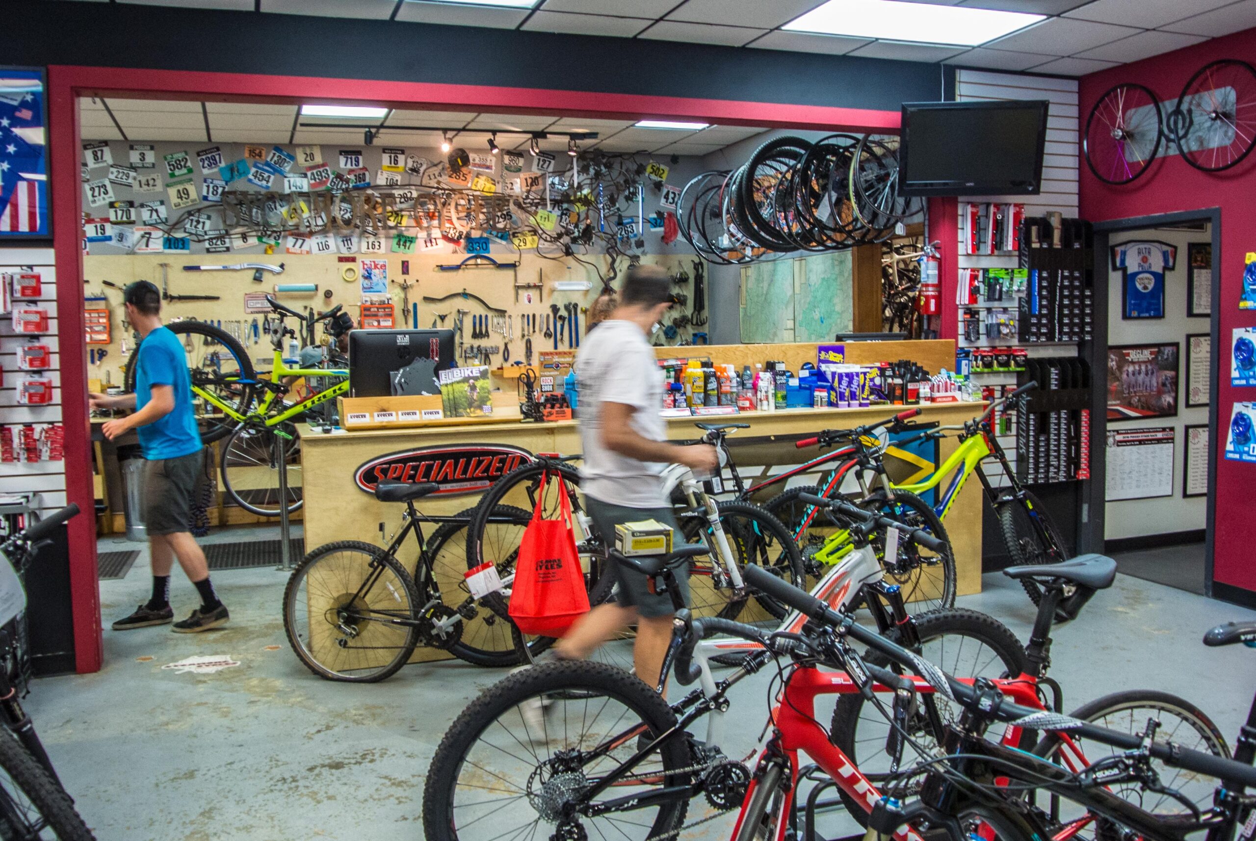 Image of a busy bike shop interior showcasing various bicycles for sale, bike accessories, and tools. Two employees are present, one working at the sales counter and the other moving around the shop. The walls are adorned with cycling memorabilia and tools, while several bike frames hang overhead.