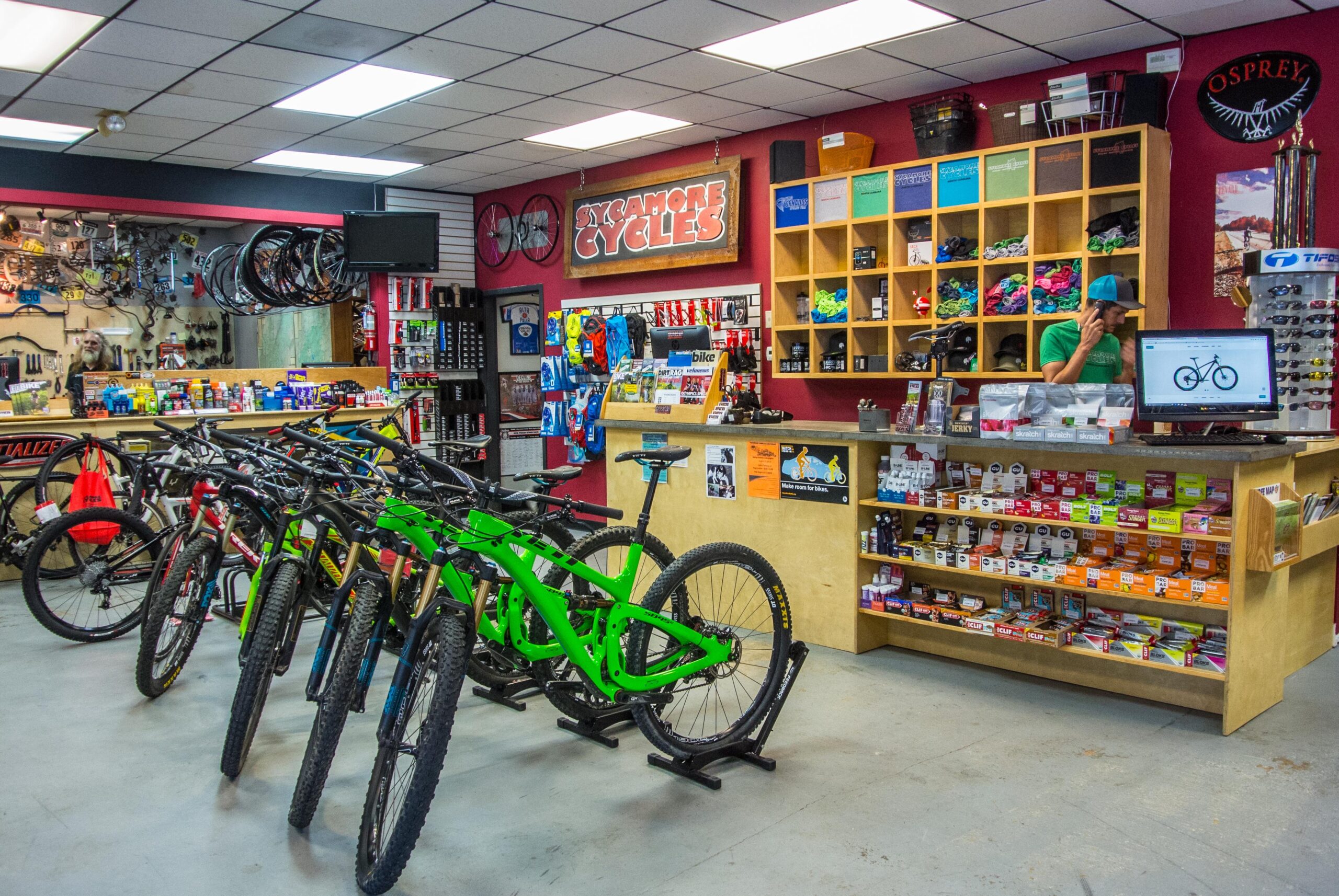 A bustling bicycle shop interior featuring a row of bikes, including brightly colored green models. The shop has a wooden counter with various cycling accessories and nutrition products displayed. Walls are adorned with bike parts, tools, and promotional materials. A staff member is engaged in a phone call at the register, and additional cycling gear is visible on shelves.
