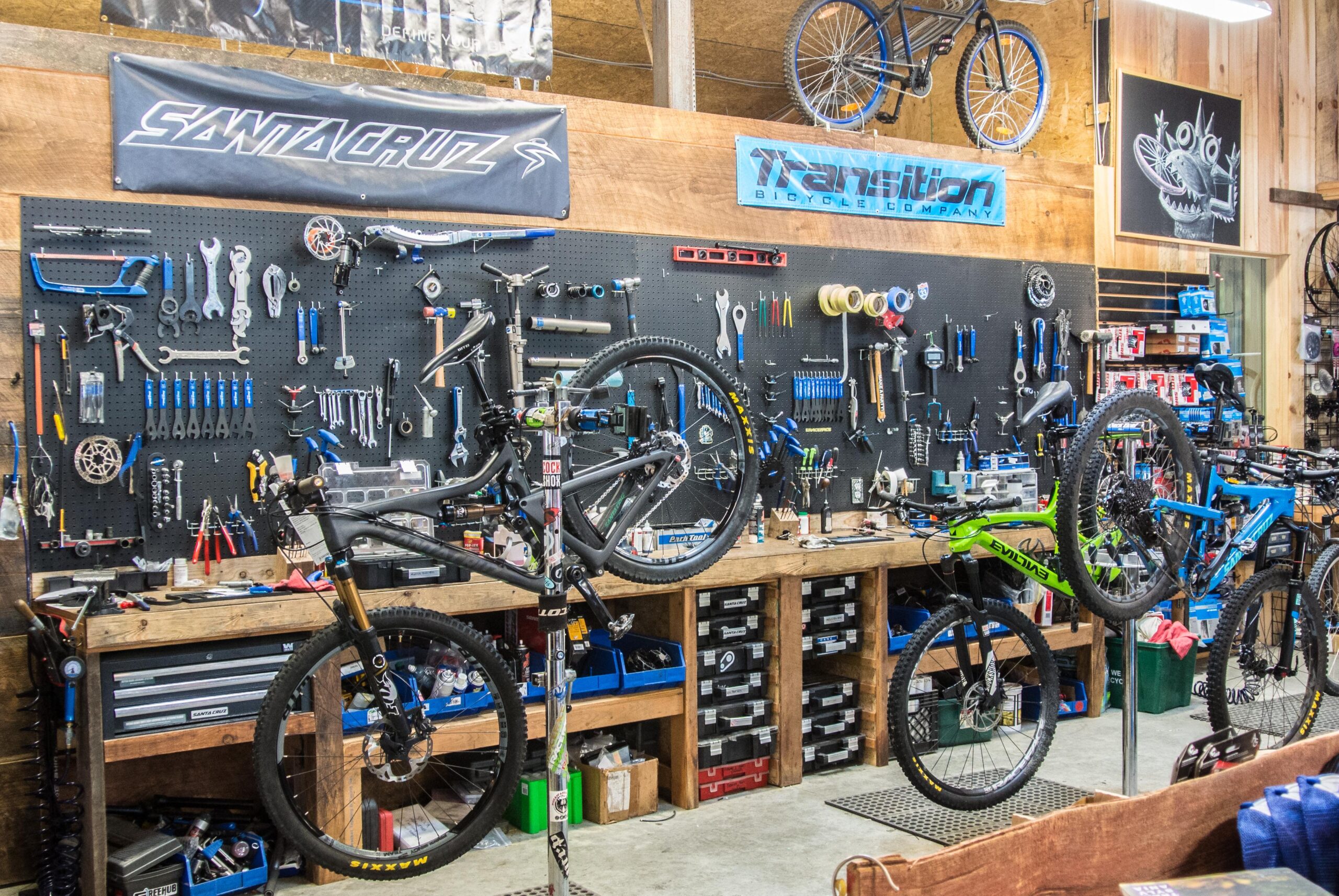 A bike repair workshop with a pegboard wall displaying various tools and equipment. Two bicycles are mounted on repair stands in the foreground, one black and one green. The workbench beneath the bikes is cluttered with toolboxes and bike parts. Banners for Santa Cruz and Transition Bicycle Company hang above the tools.