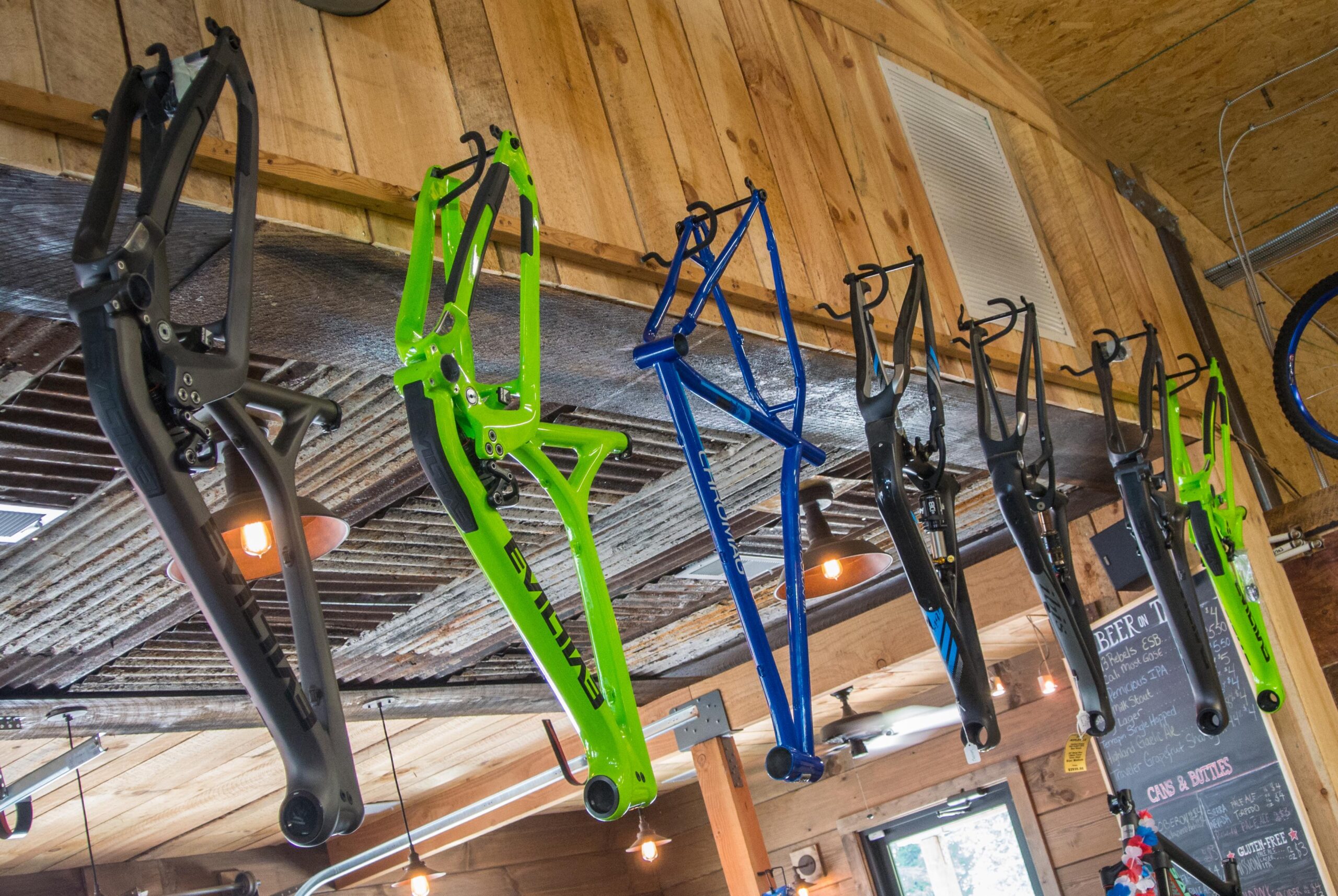 Alt text: A row of bicycle frames in various colors, including black, green, and blue, hanging from hooks in a rustic interior with wooden walls and soft lighting.
