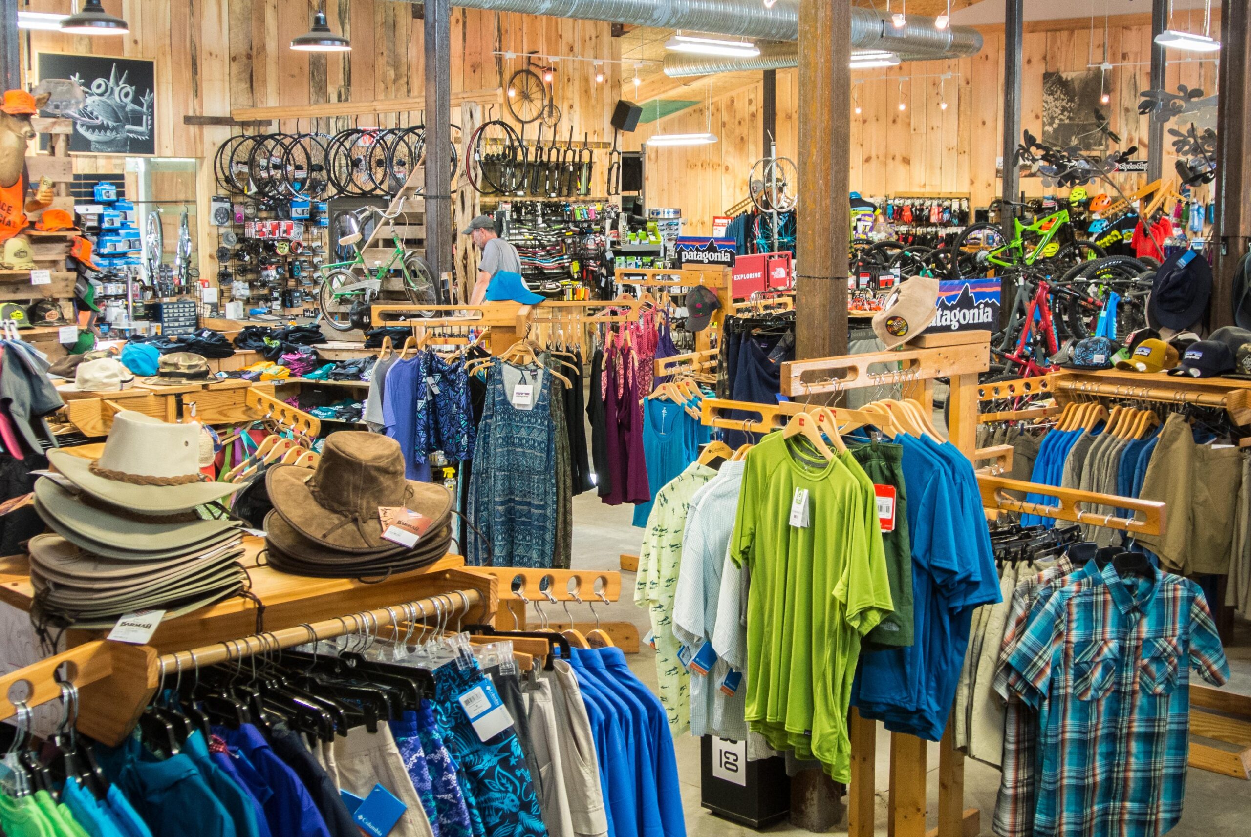 Image of an outdoor gear store featuring a wide assortment of clothing, hats, and cycling equipment. Rows of colorful shirts, shorts, and hats are displayed on wooden racks in the foreground, while bicycles and other sporting goods are visible in the background. The store has a warm, rustic feel with wooden walls and hanging lights, creating a welcoming shopping atmosphere.