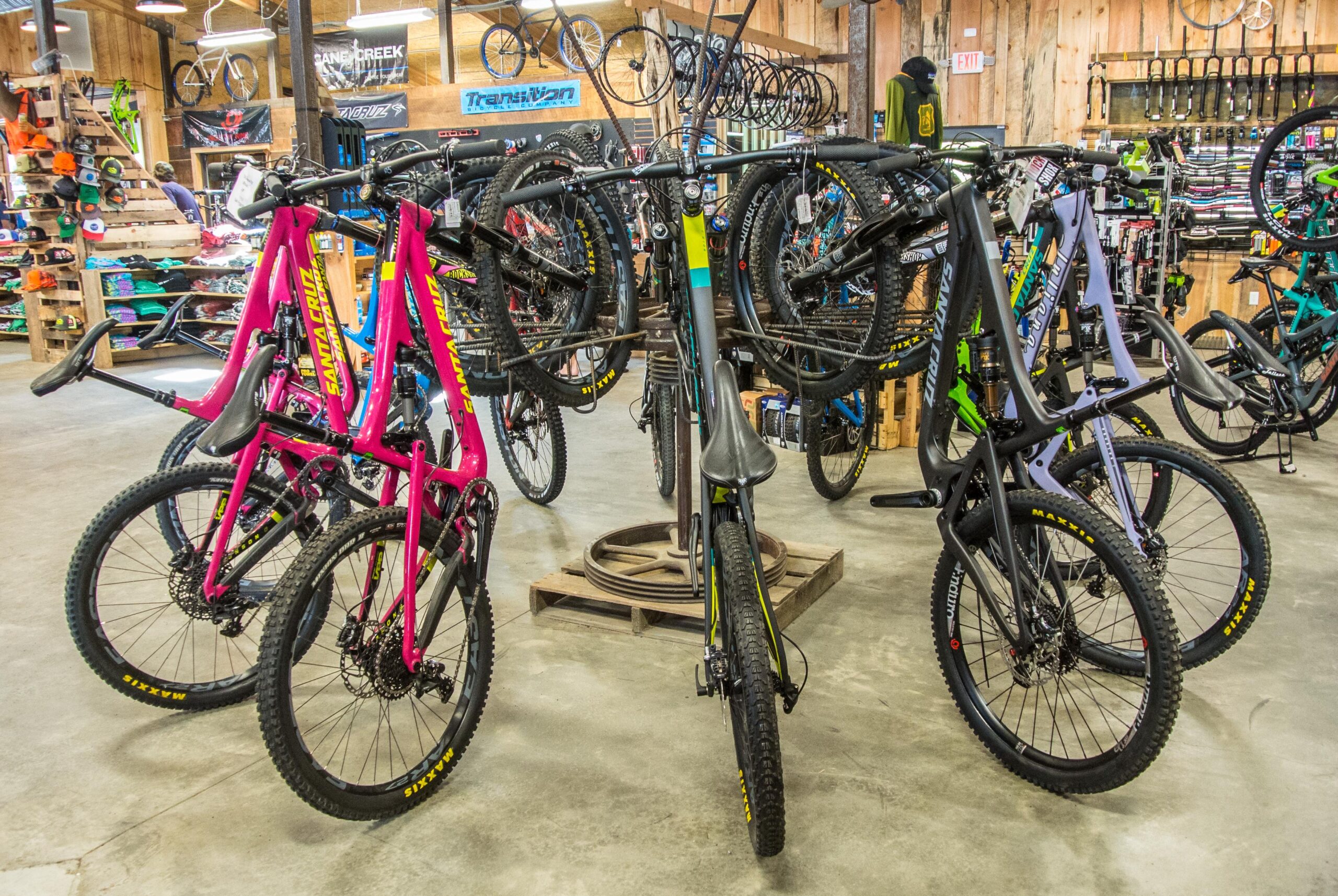 A display of various mountain bikes in a bike shop, featuring brightly colored frames, including pink and black. The bikes are arranged on a central rack, with additional bike accessories visible on the shelves in the background. The shop has a rustic wooden interior, enhancing the outdoor sporting vibe.
