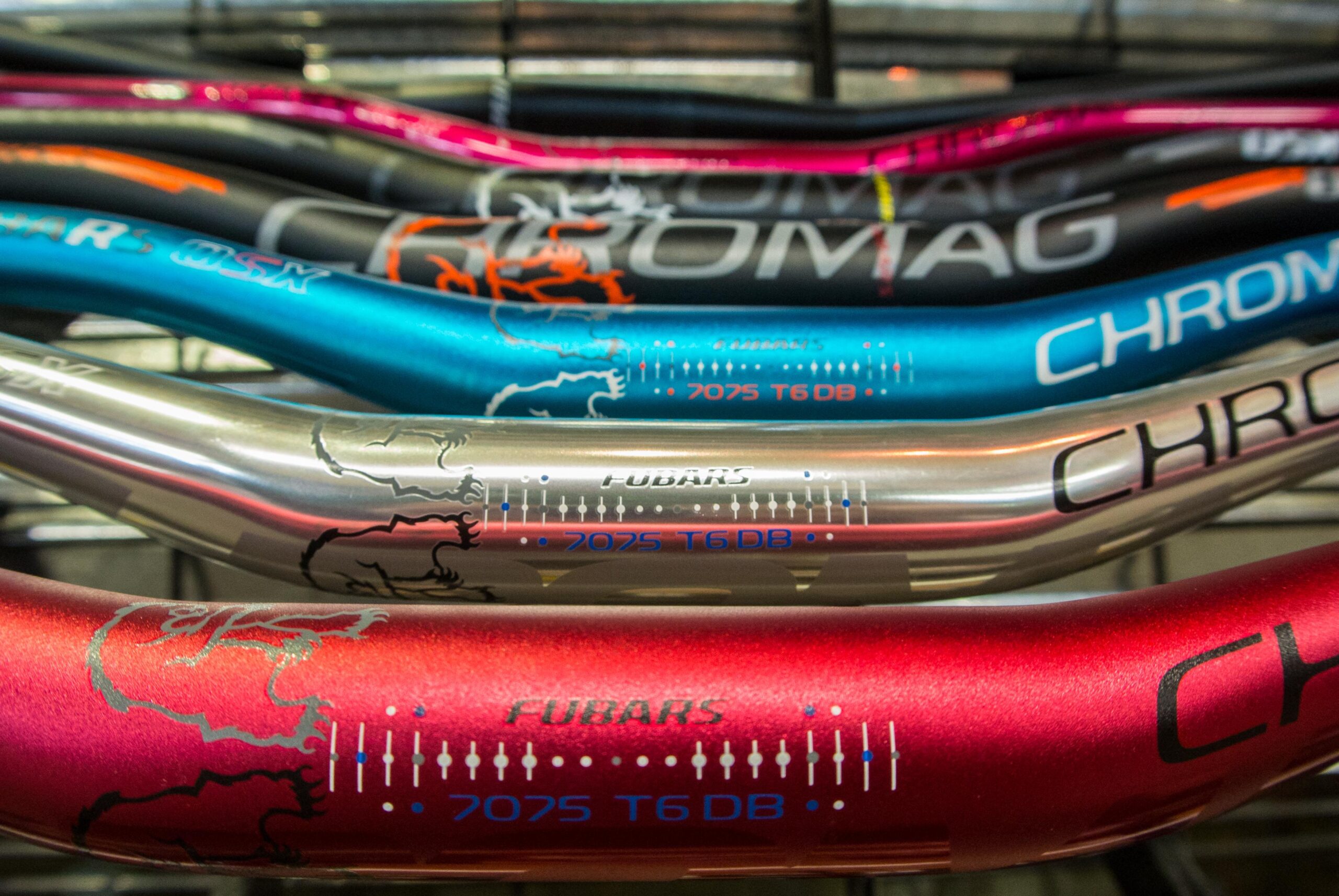 Photo of various colorful bike handlebars displayed in a row. The handlebars feature different colors, including pink, blue, silver, and red, with visible branding and specifications, such as "7075 T6 DB." The design showcases a mix of matte and shiny finishes, highlighting their sporty appearance.