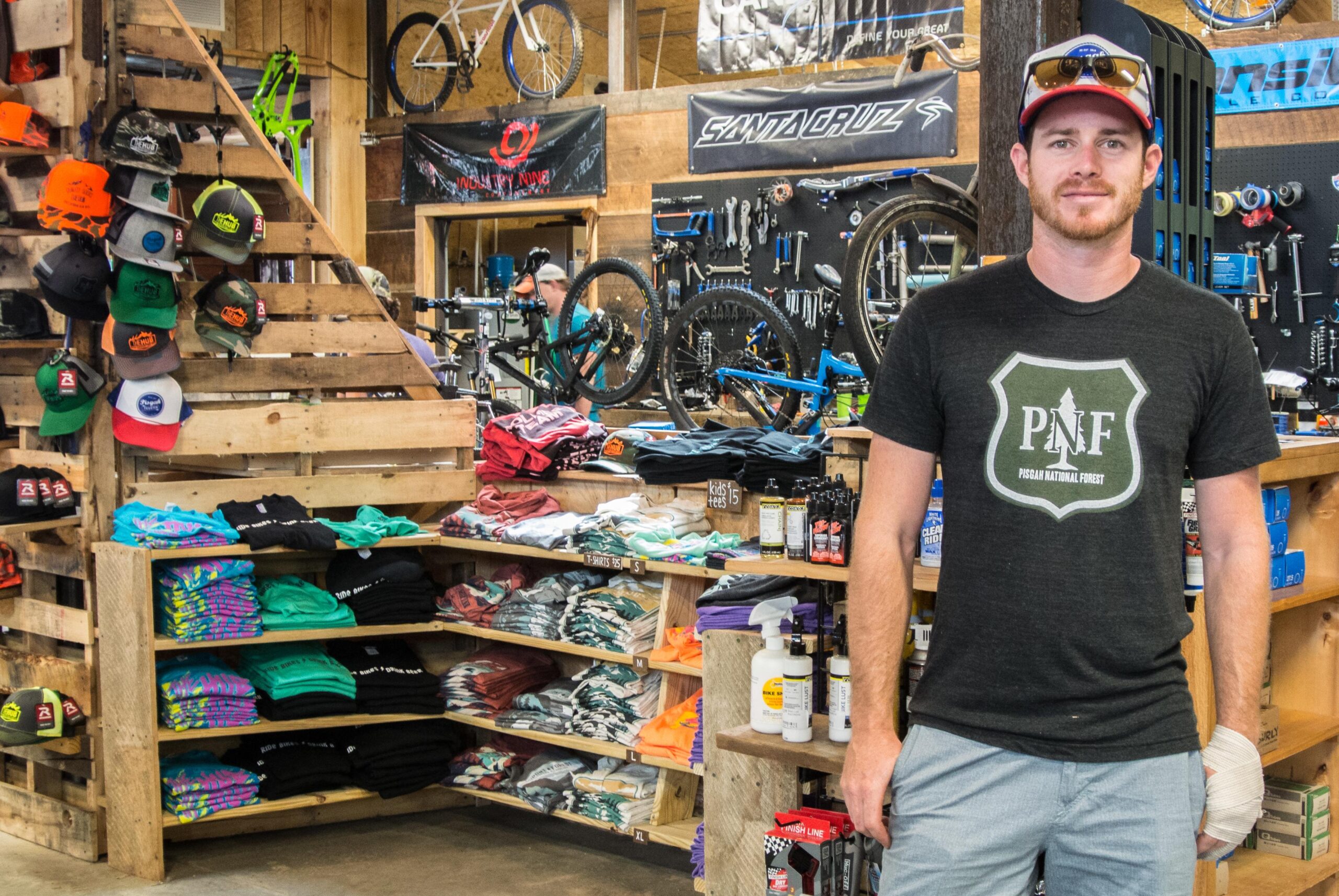 A man stands in a bicycle shop surrounded by colorful T-shirts, hats, and bike accessories. He wears a dark shirt featuring a logo for Pisgah National Forest and is smiling at the camera. In the background, various bicycles and tools are displayed.