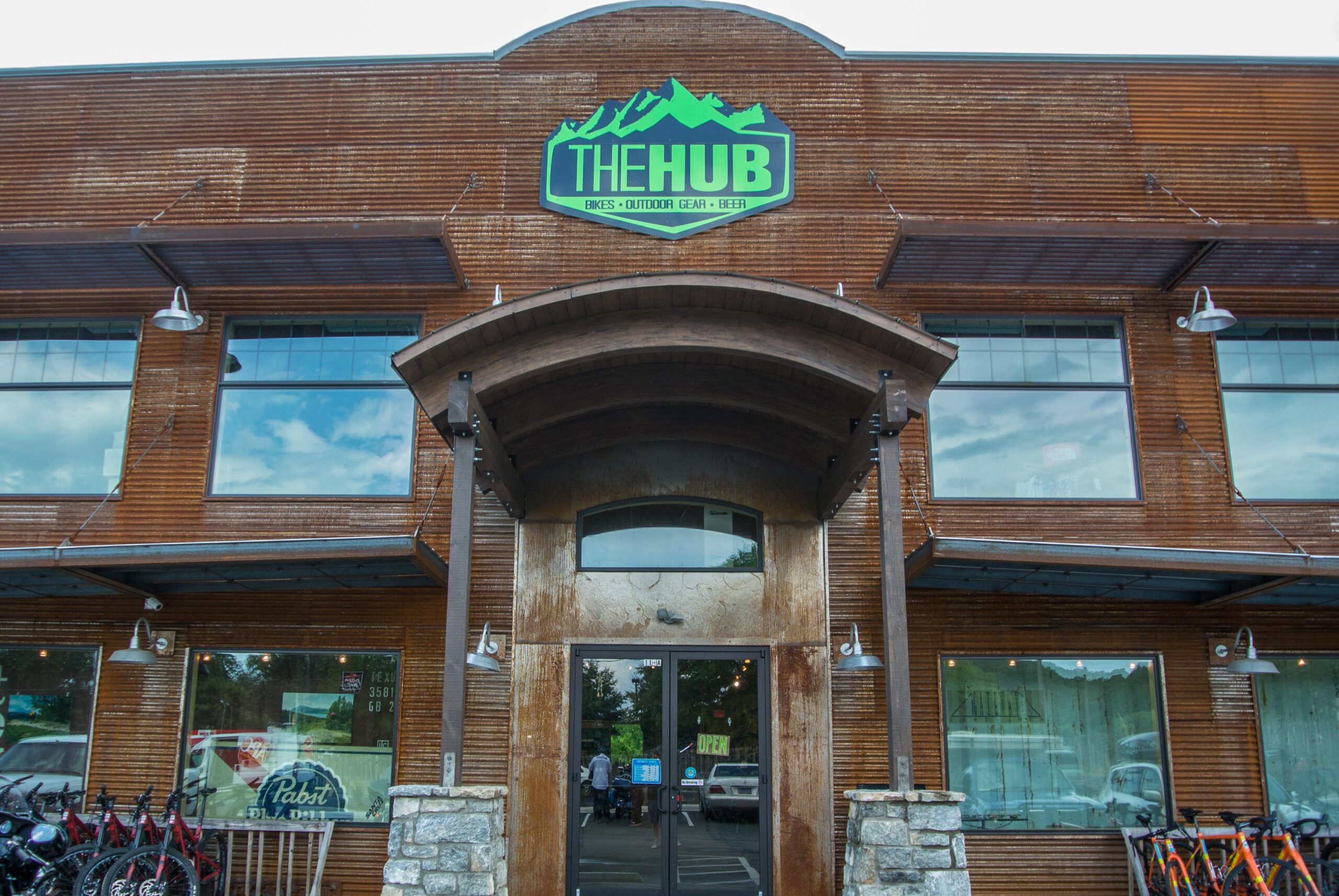 Exterior view of a building named "The Hub," featuring a wooden facade and large glass windows. The entrance has a curved roof and a wooden porch. Bicycles are parked outside, and a sign indicates that the shop offers bikes, outdoor gear, and beer. The atmosphere suggests a welcoming hub for outdoor enthusiasts.