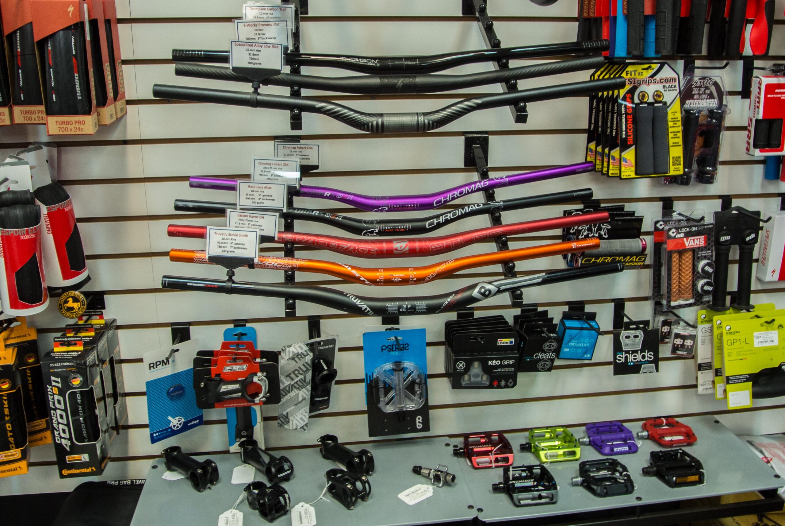 A display of various bicycle handlebars and accessories, including colorful handlebars in purple, orange, and black, along with pedals, grips, and other cycling components. The items are organized on a wall-mounted rack, with price tags and product information visible.