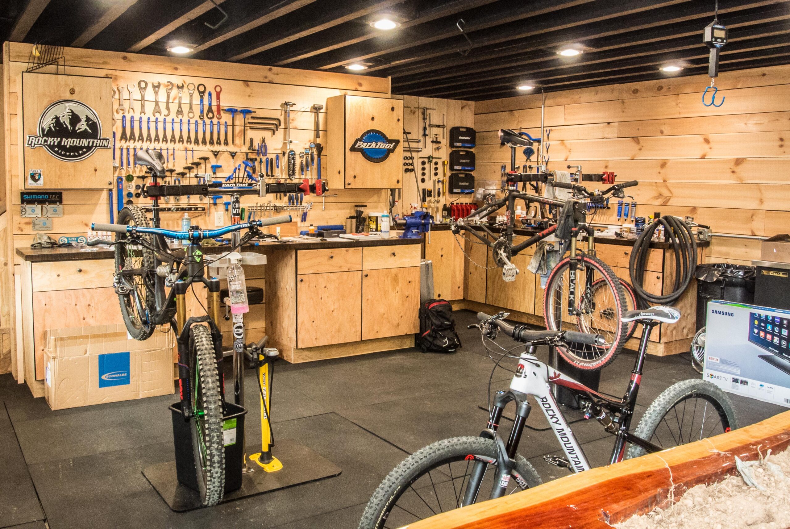 An interior view of a well-equipped bike workshop featuring two bicycles on repair stands, surrounded by a variety of tools organized on wooden shelves. The space includes a workbench with additional tools and equipment, a television in the corner, and a clean, organized environment with wooden paneling and rubber flooring.
