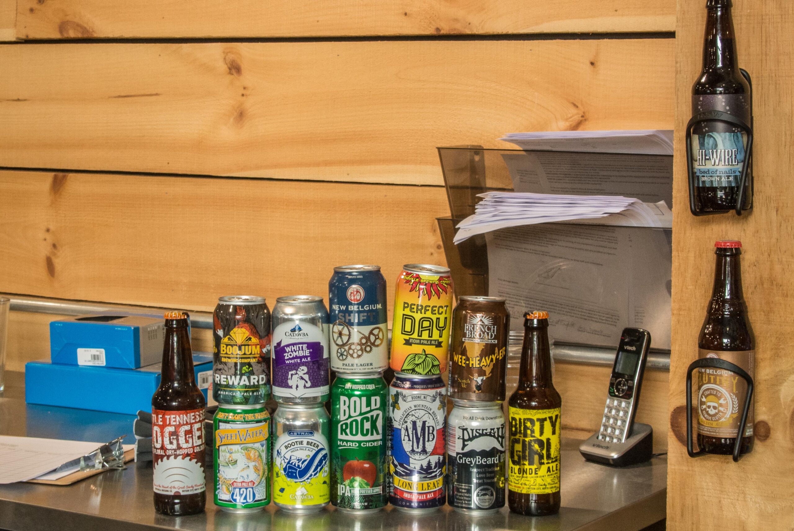 A collection of various beer cans and bottles arranged on a countertop, featuring brands like New Belgium, Bold Rock, and Hi-Wire. Behind the cans, there is a wooden wall and a small stack of papers. A telephone sits on the countertop beside the beverages.