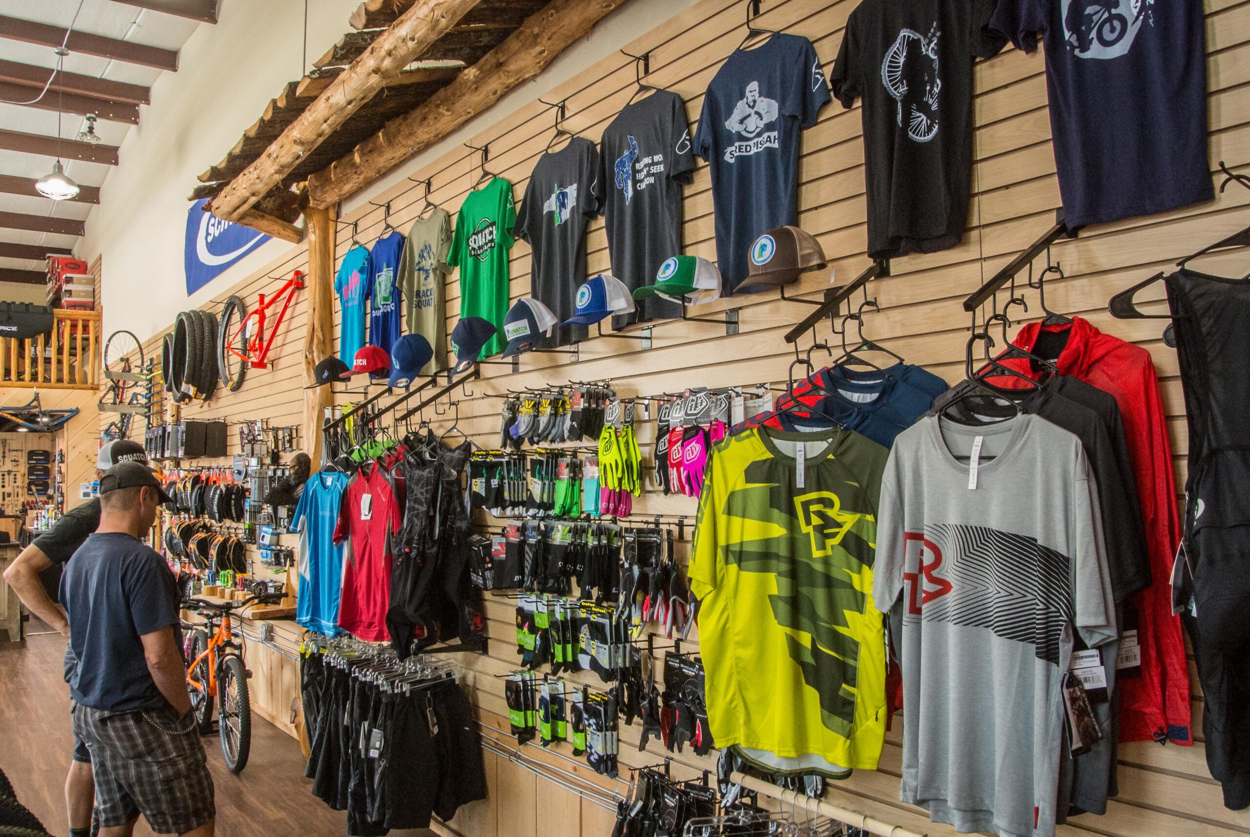 A brightly lit bike shop interior featuring a wooden wall display of various cycling apparel, including t-shirts, hats, and gloves. Two customers are browsing the selection, which includes colorful shirts and accessories hanging on racks. The background displays bike parts and accessories, enhancing the atmosphere of a dedicated cycling retail space.