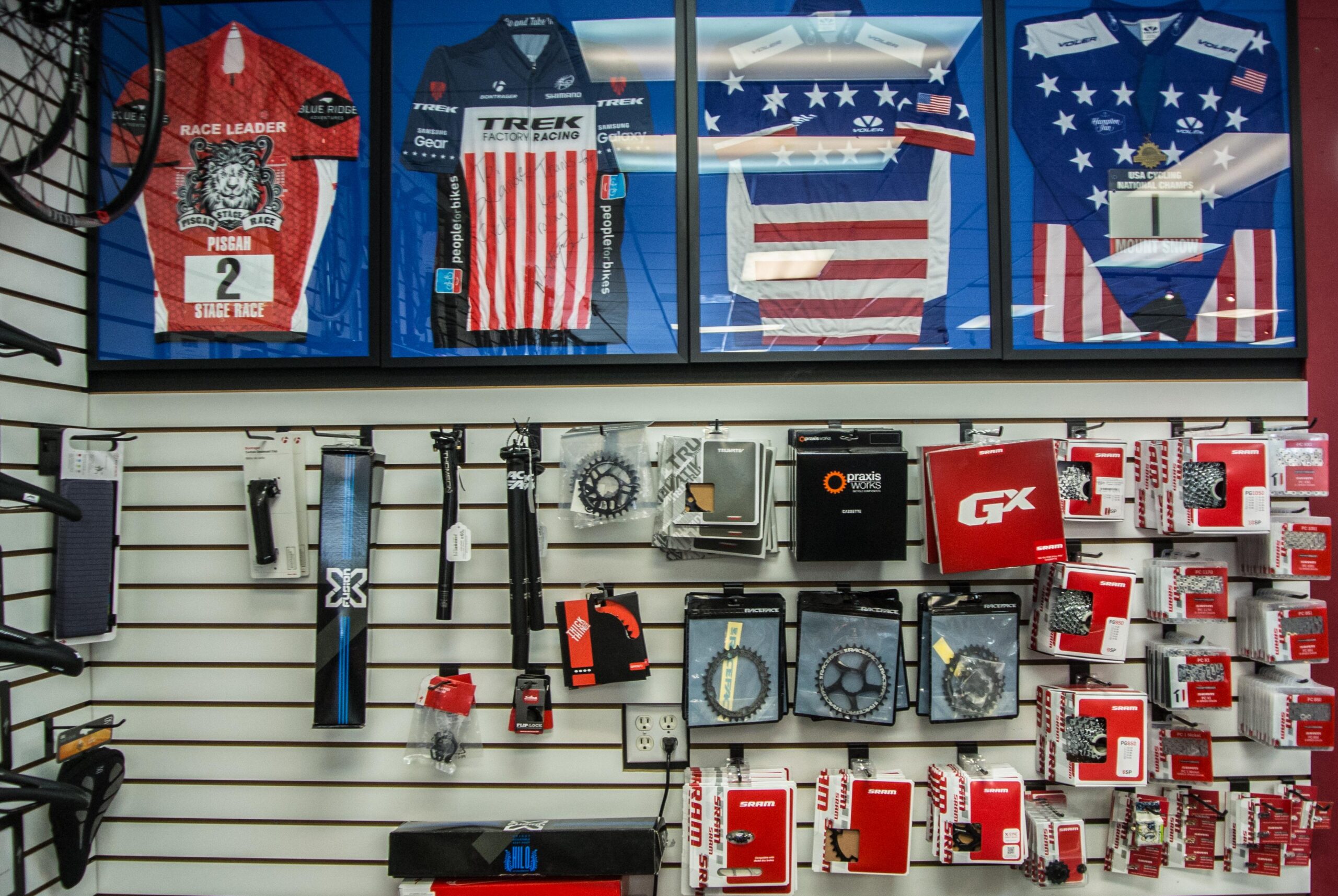 A display wall featuring framed cycling jerseys, including a red "Race Leader" jersey and two jerseys with American flag designs, alongside a variety of bike components and accessories arranged on hooks.