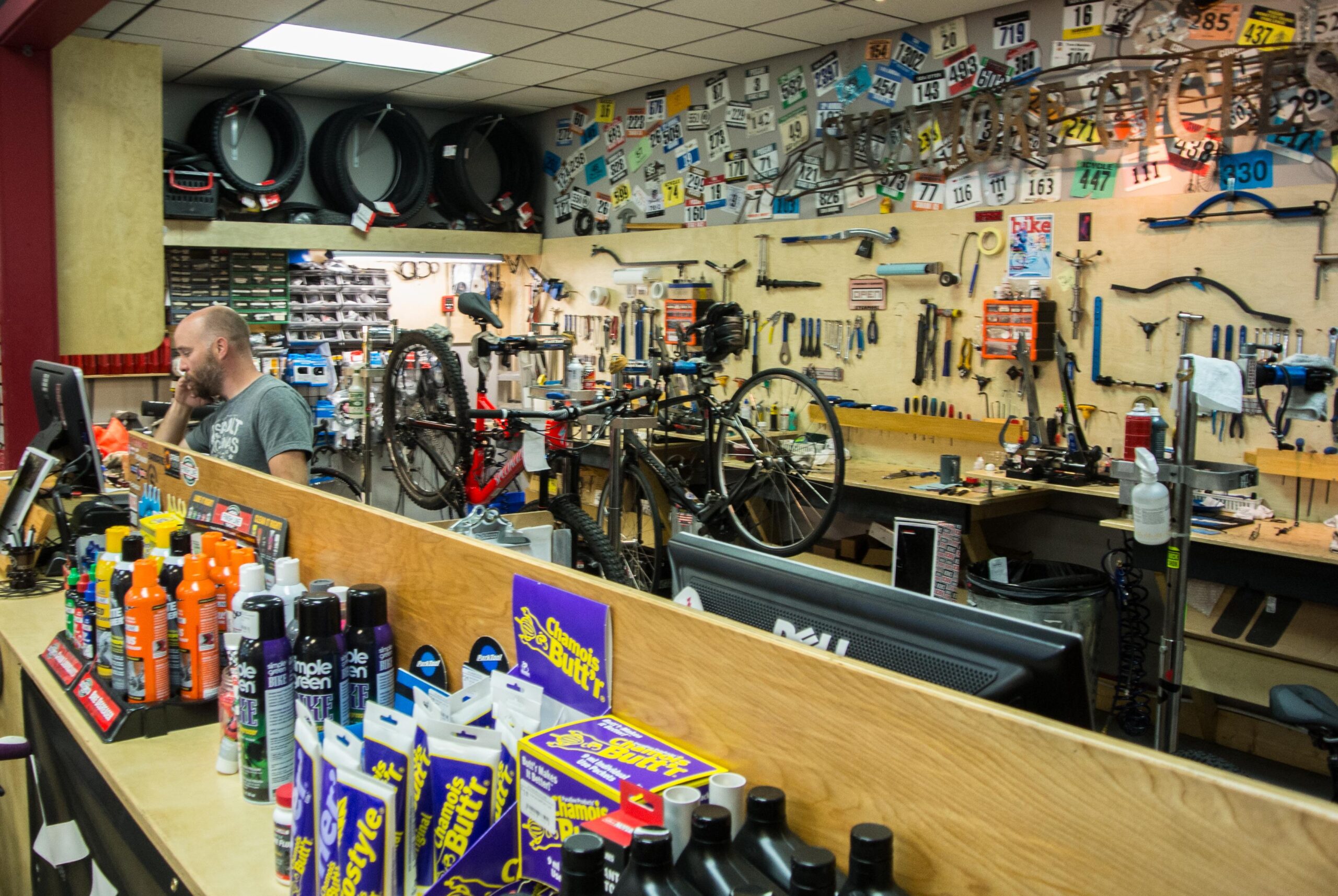 A bicycle shop interior featuring a workshop area with tools mounted on the wall and two bicycles on repair stands. A staff member is seated at the counter, speaking on the phone, while various bike maintenance products, including cleaners and lubricants, are displayed on the counter. Bike tires are stacked on the wall in the background, creating a busy but organized atmosphere.
