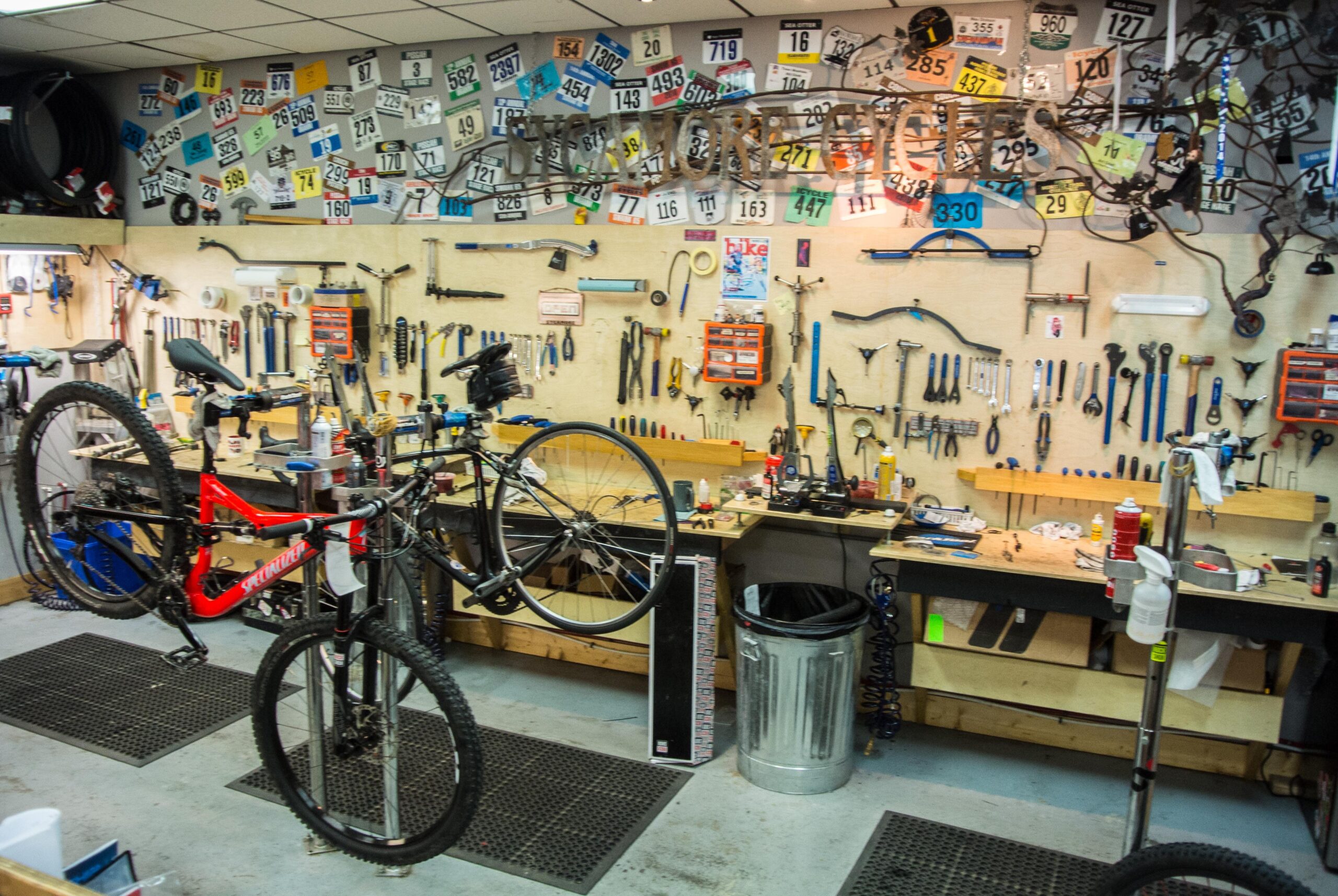 A bike repair workshop featuring two bicycles on stands, surrounded by various tools and equipment. The walls are adorned with an array of race numbers and cycling memorabilia, while the workbenches are cluttered with tools, repair items, and a trash can. The workspace has a functional and organized vibe, suitable for bicycle maintenance and repairs.