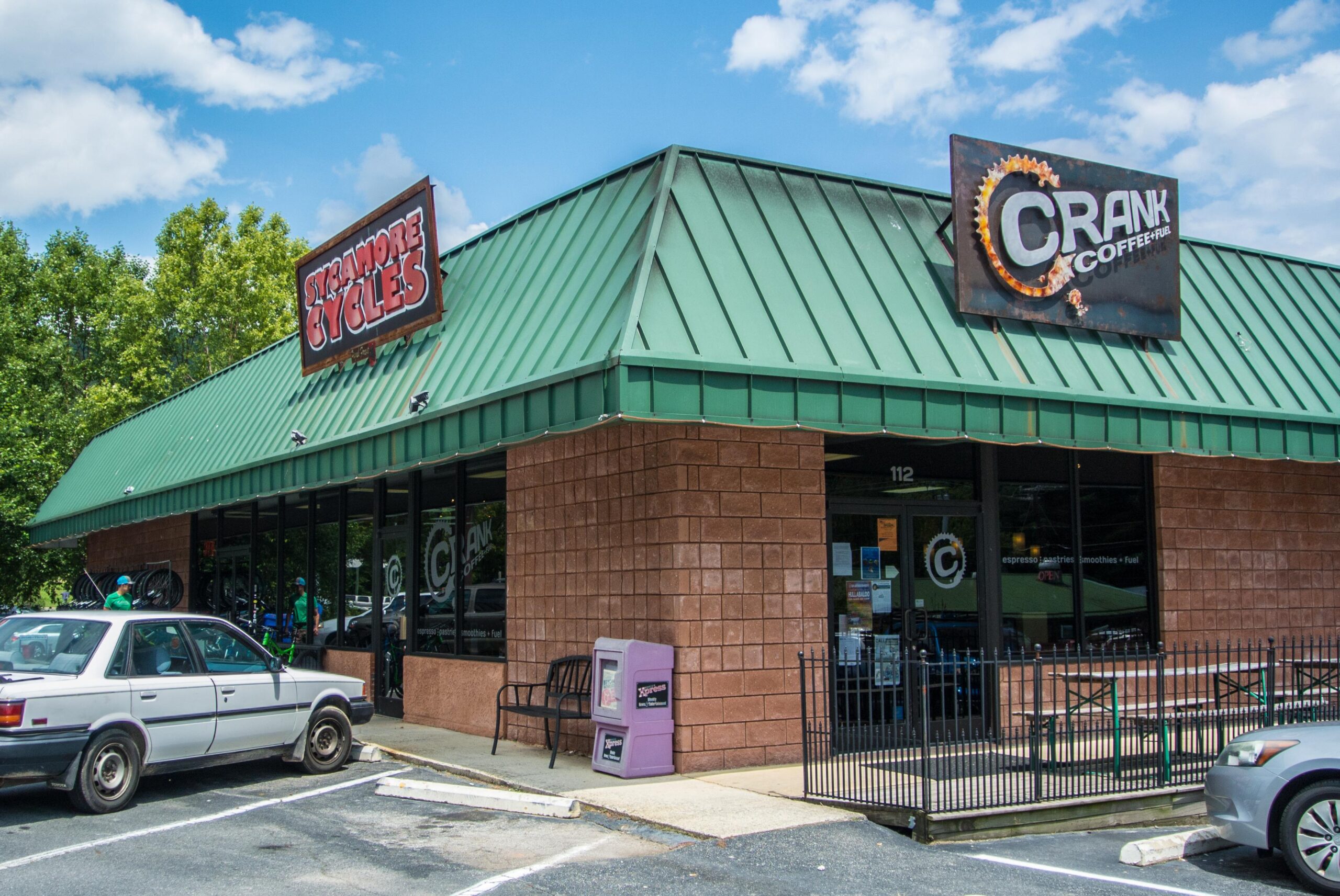 Image of a building with a green metal roof featuring two signs: one for "Stramore Cycles" and another for "Crank Coffee". The storefront has large windows displaying bicycles and seating outside. A white car is parked in front, and the area is surrounded by trees under a partly cloudy blue sky.