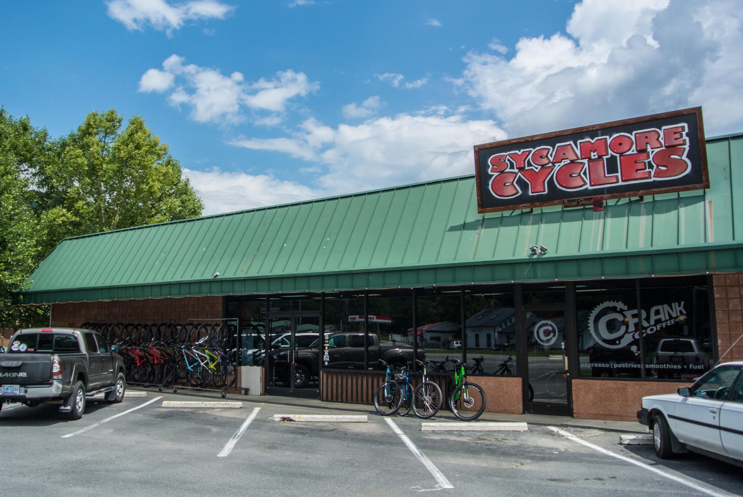 Alt text: A storefront of Sycamore Cycles featuring a green metal roof and a large sign above. Bicycles are displayed outside, and there are several parked cars in front of the shop. The sky is partly cloudy.