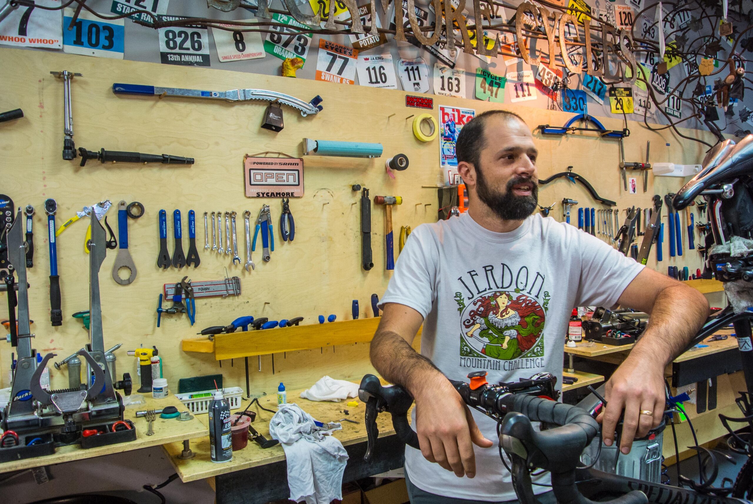 A man with a beard wearing a white T-shirt stands near a bicycle in a workshop filled with various tools. Behind him is a wall adorned with bike-related memorabilia and a variety of tools organized neatly. The workspace has a professional yet inviting atmosphere, highlighting a passion for cycling and bike maintenance.
