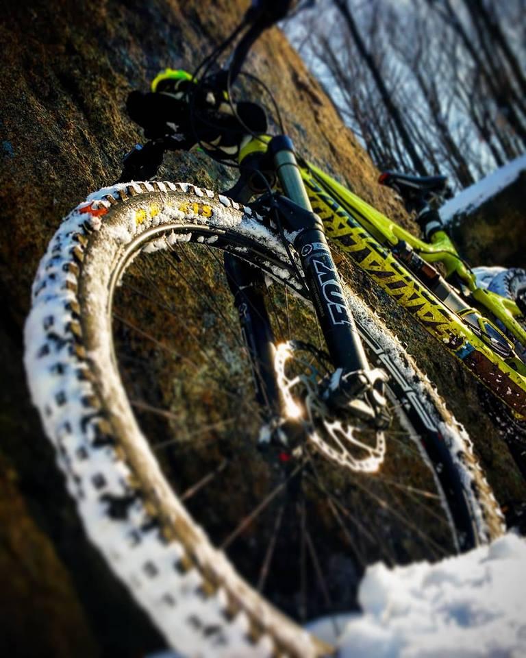 Santa Cruz Bronson: A close-up image of a mountain bike leaning against a rock, with snow covering the tire. The bike features a vibrant green frame and specialized tires designed for winter conditions. The background shows a blurred winter landscape.