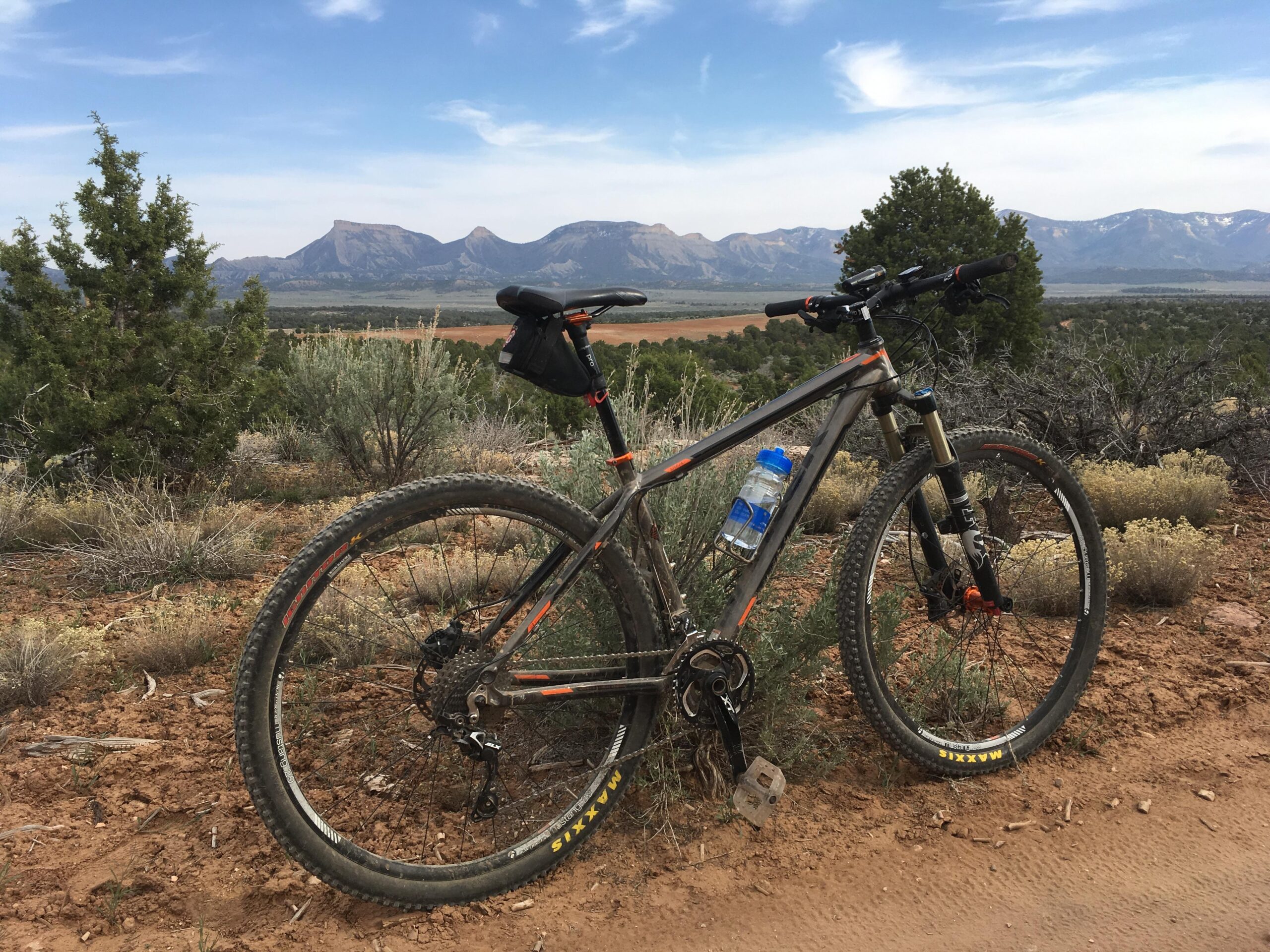 Trek Superfly: A mountain bike rests on a dirt trail surrounded by shrubs and low vegetation, with a backdrop of rugged mountains under a blue sky. A water bottle is attached to the bike, emphasizing an outdoor adventure setting.
