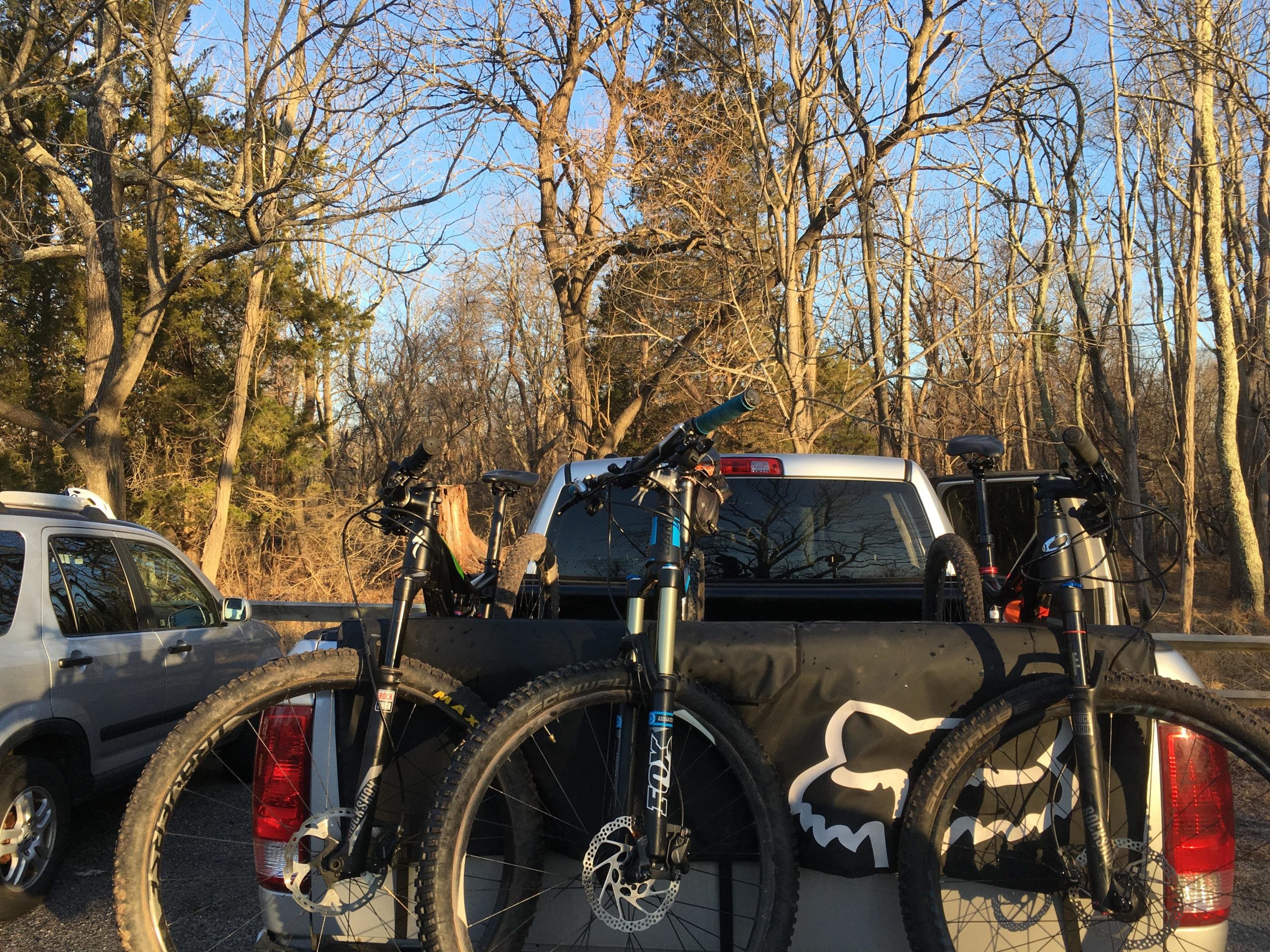 A view of the back of a pickup truck with two mountain bikes secured in the truck bed. The surrounding area features bare trees indicating early spring or late fall, with a clear blue sky overhead. Another vehicle is parked nearby, and the scene is set in a natural environment. Allaire State Park mountain bike trail.