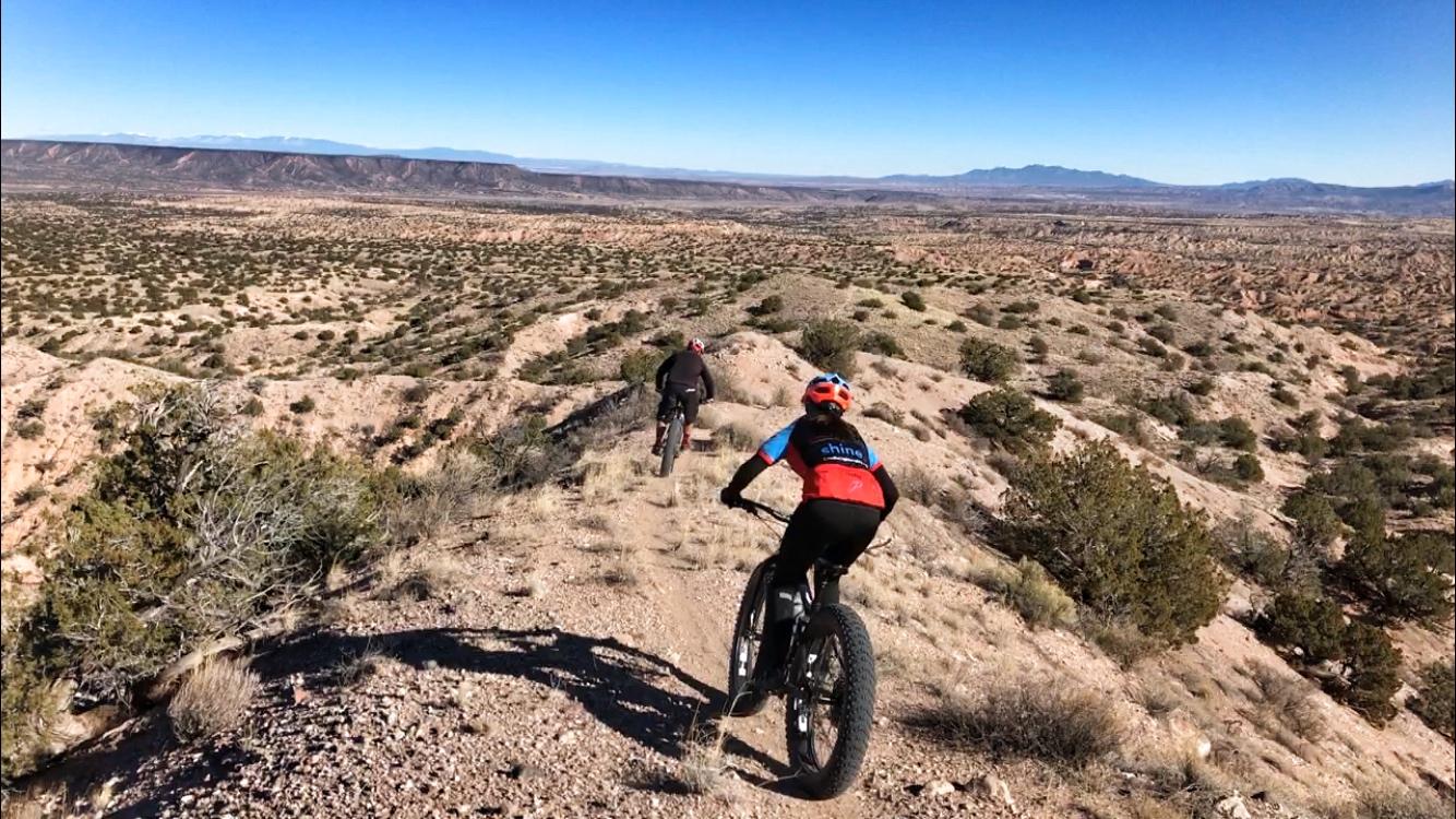 Two mountain bikers riding along a rocky trail in a desert landscape, with shrubs and sparse vegetation. The terrain features hills and distant mountains under a clear blue sky. Mariposa Fat Bike Trails mountain bike trail.