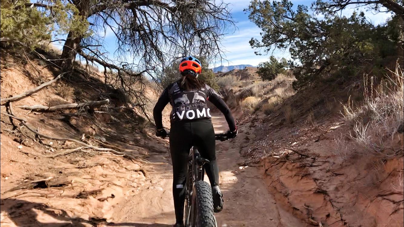A person riding a mountain bike on a dirt trail surrounded by sparse trees and shrubs under a clear blue sky. The rider is wearing a helmet and cycling gear, and the path appears winding and rugged. Mariposa Fat Bike Trails mountain bike trail.