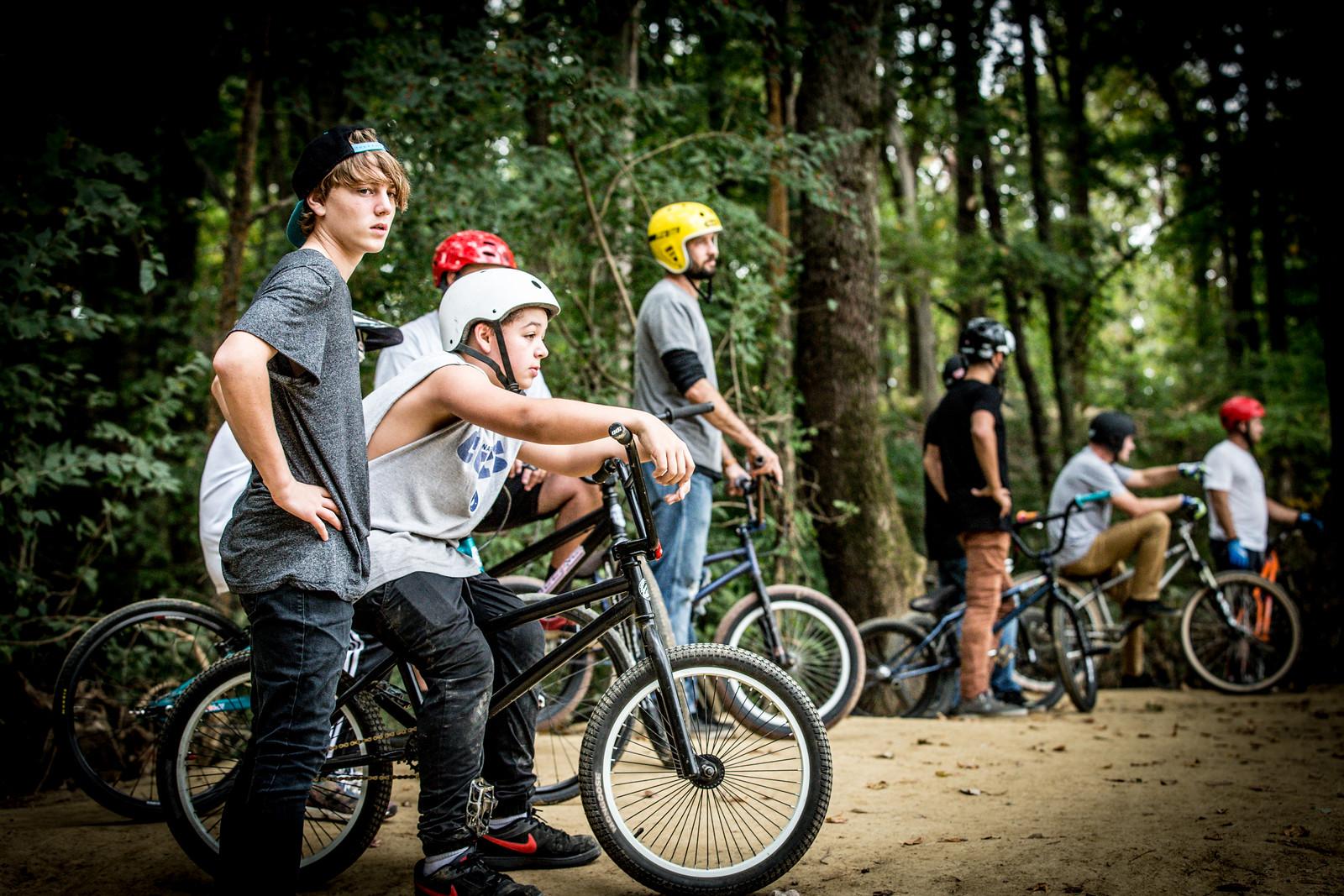 A group of young riders during a BMX session in a wooded area. Two boys in the foreground are seated on their bikes, wearing helmets, while others stand nearby, also on their bikes. The scene captures an active and adventurous atmosphere. Griffin Bike Park mountain bike trail.