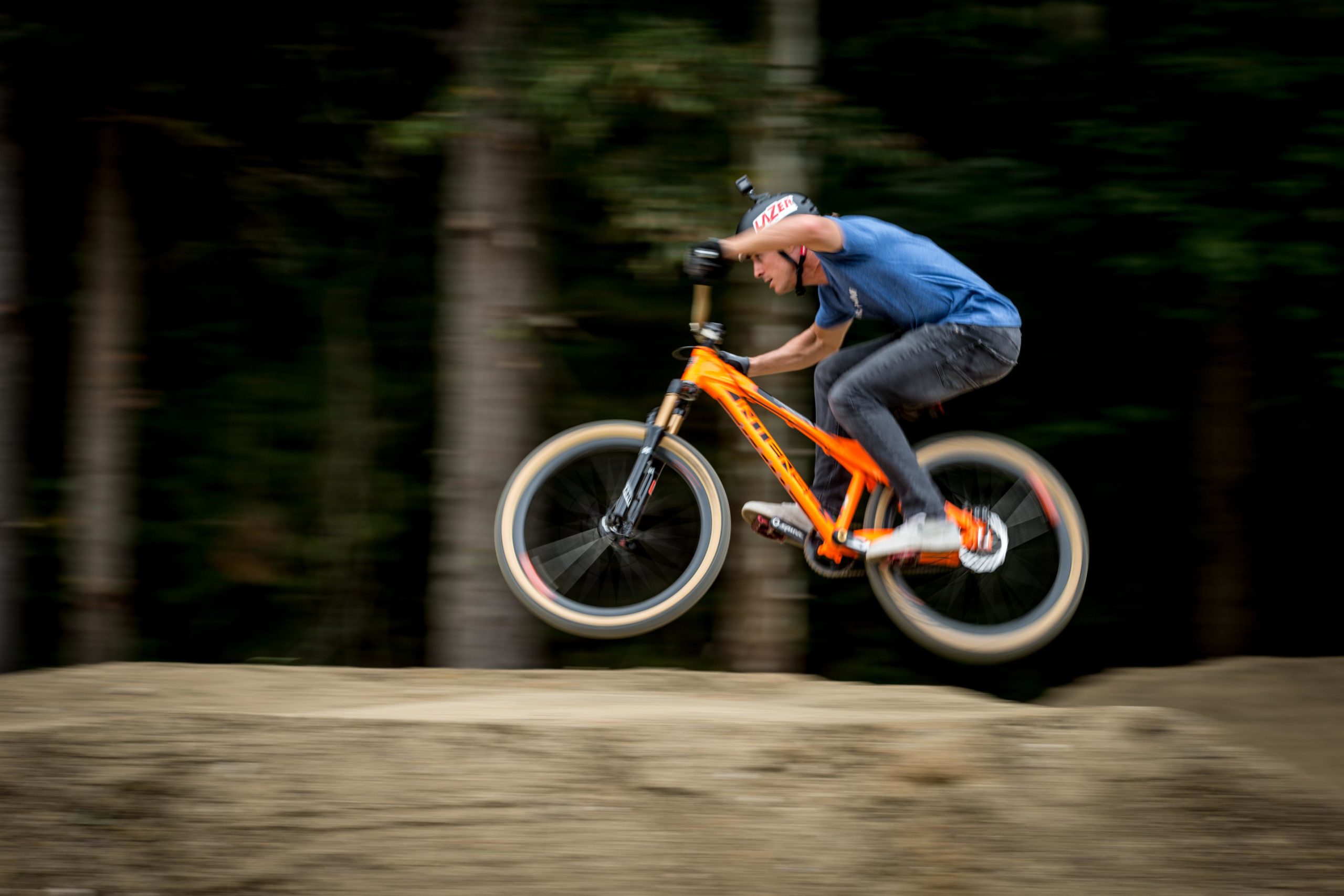 A cyclist performing a jump on an orange mountain bike over a dirt ramp, captured in motion against a blurred forest background. Griffin Bike Park mountain bike trail.