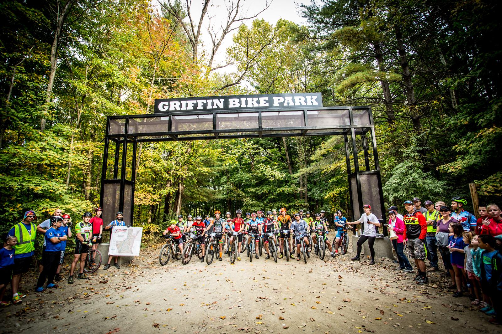 A group of cyclists and spectators gather at the entrance of Griffin Bike Park, marked by a prominent sign. The scene features lush greenery with autumn foliage, and participants are lined up on bikes, some holding a map. The atmosphere is lively and anticipatory as it appears to be the start of an event. Griffin Bike Park mountain bike trail.