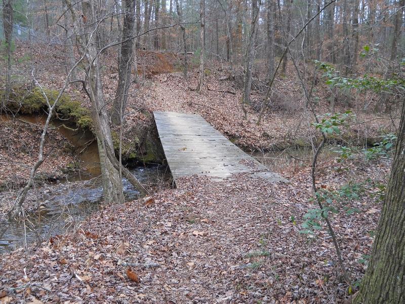 A wooden bridge spans a small creek in a wooded area, surrounded by fallen leaves and bare trees. The path leading up to the bridge is covered in autumn foliage, creating a tranquil natural landscape. Etowah River Trail mountain bike trail.