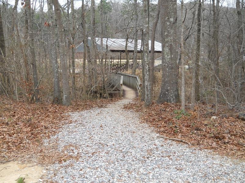 A gravel path leads through a forested area with bare trees, guiding toward a wooden bridge that connects to a building with a metal roof in the background. The ground is covered with fallen leaves, and the scene conveys a quiet, natural setting. Montgomery Creek Trail mountain bike trail.