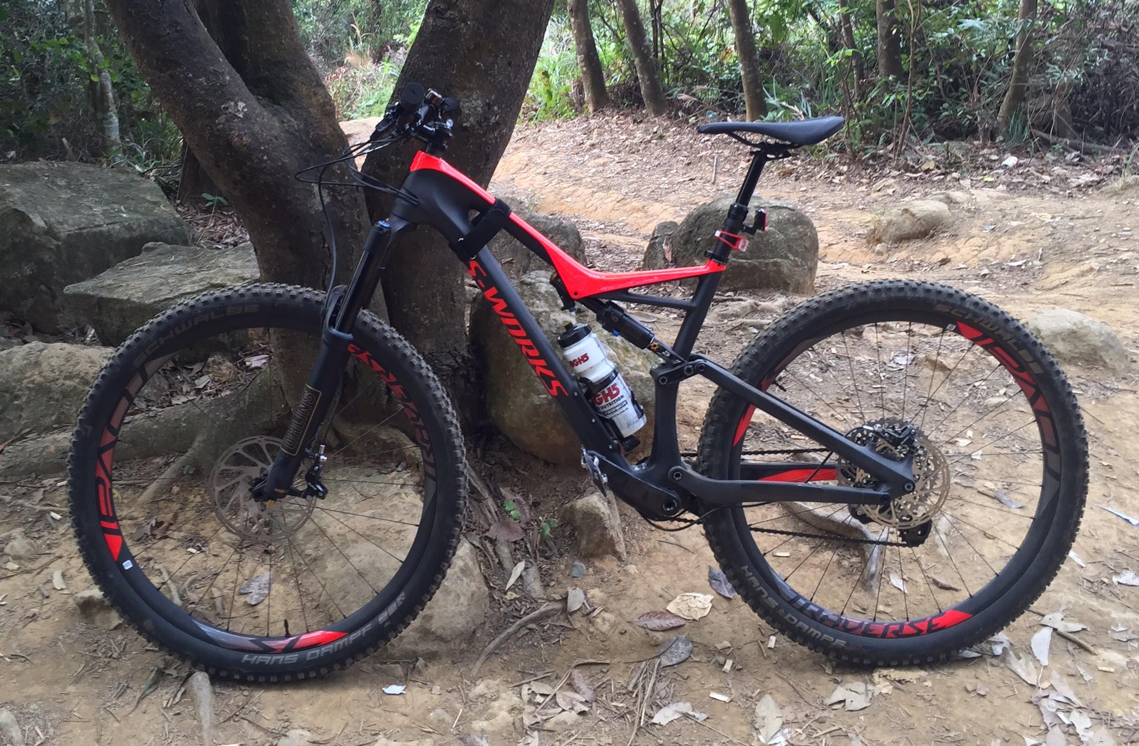 Specialized Stumpjumper S Works FSR: A mountain bike featuring a black and red frame, positioned against a tree in a forested area. The bike has wide tires designed for off-road terrain, with a water bottle mounted on the frame, surrounded by rocks and dirt trails.