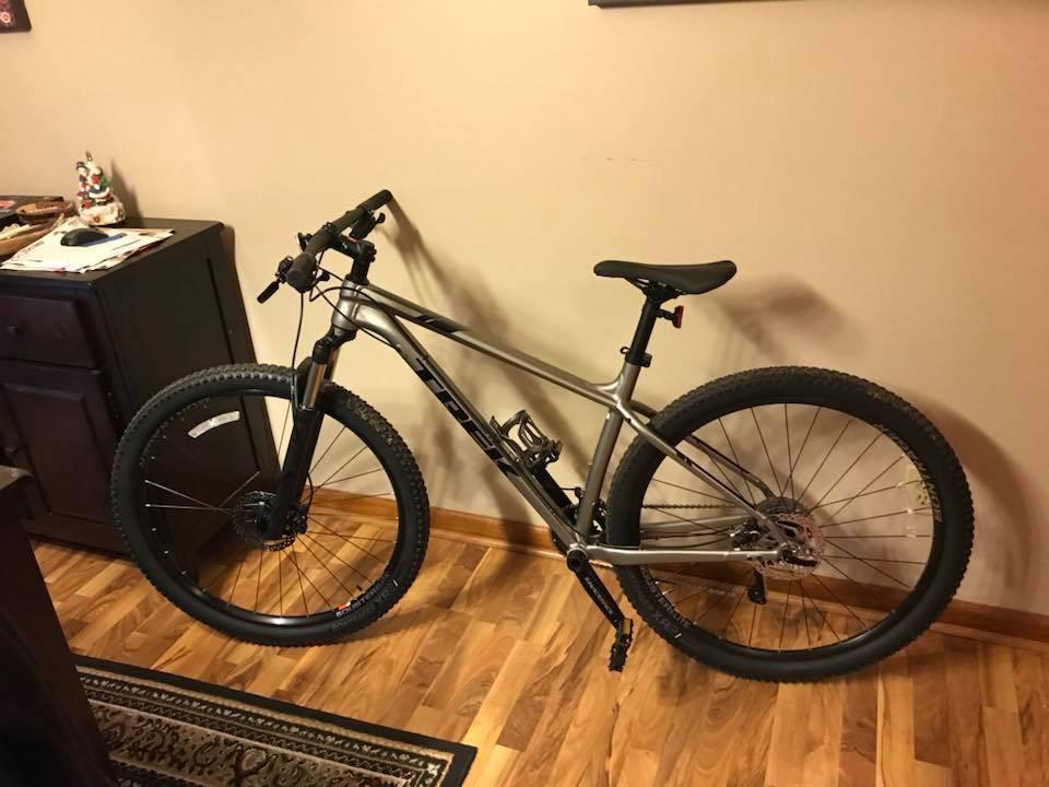 Trek Xcaliber 8: A mountain bike leaning against a wall in a home interior. The bike has a metallic gray frame, wide tires, and front suspension. In the background, there is a small cabinet with some papers and a decorative item on top, as well as wooden flooring.