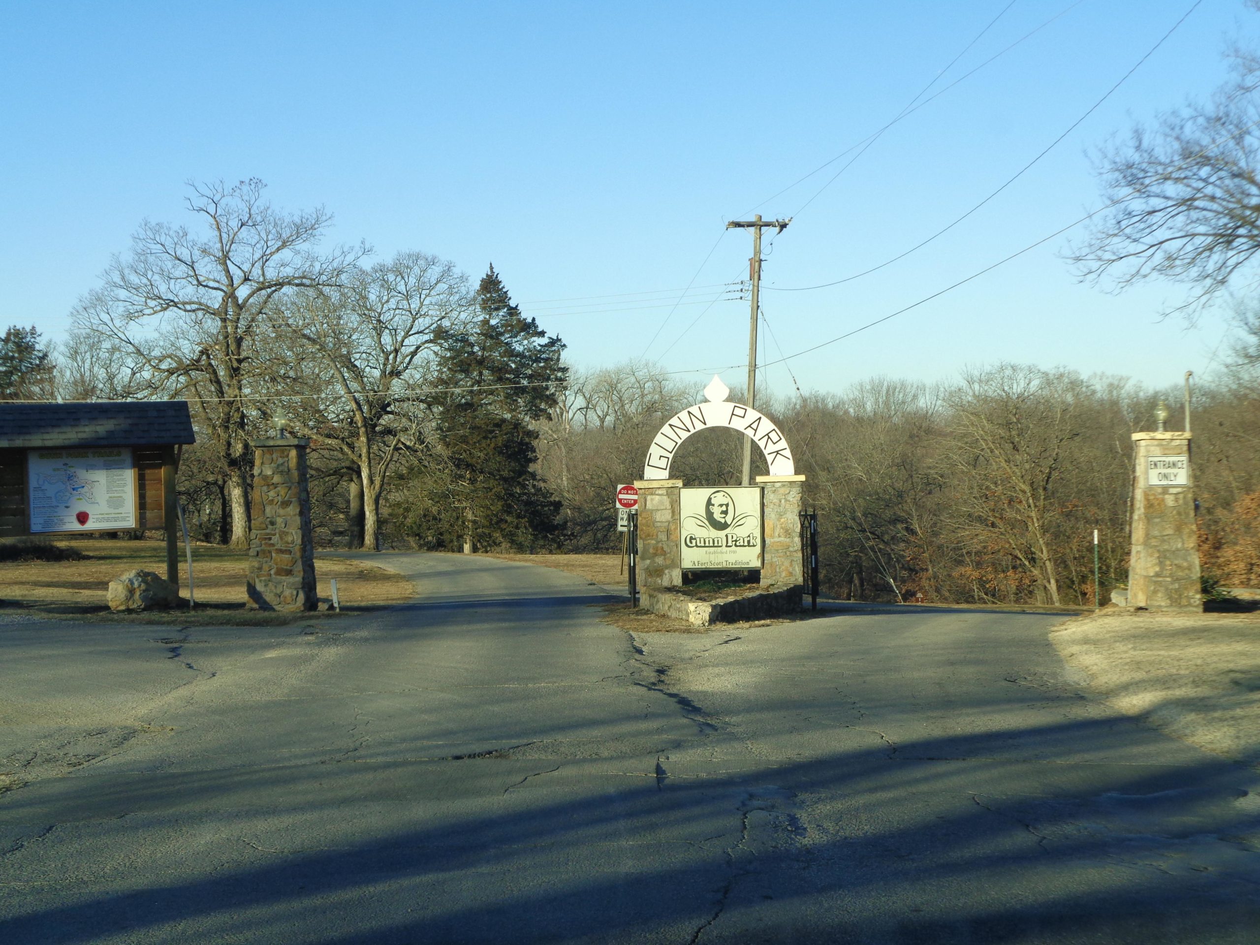 Entrance to Gunn Park, featuring a stone archway sign and surrounding trees on a clear day. A sign on the left displays a map of the park, and a "No Entry" sign is visible on the right. The road leads inward, inviting visitors into the park area. Gunn Park Trails mountain bike trail.