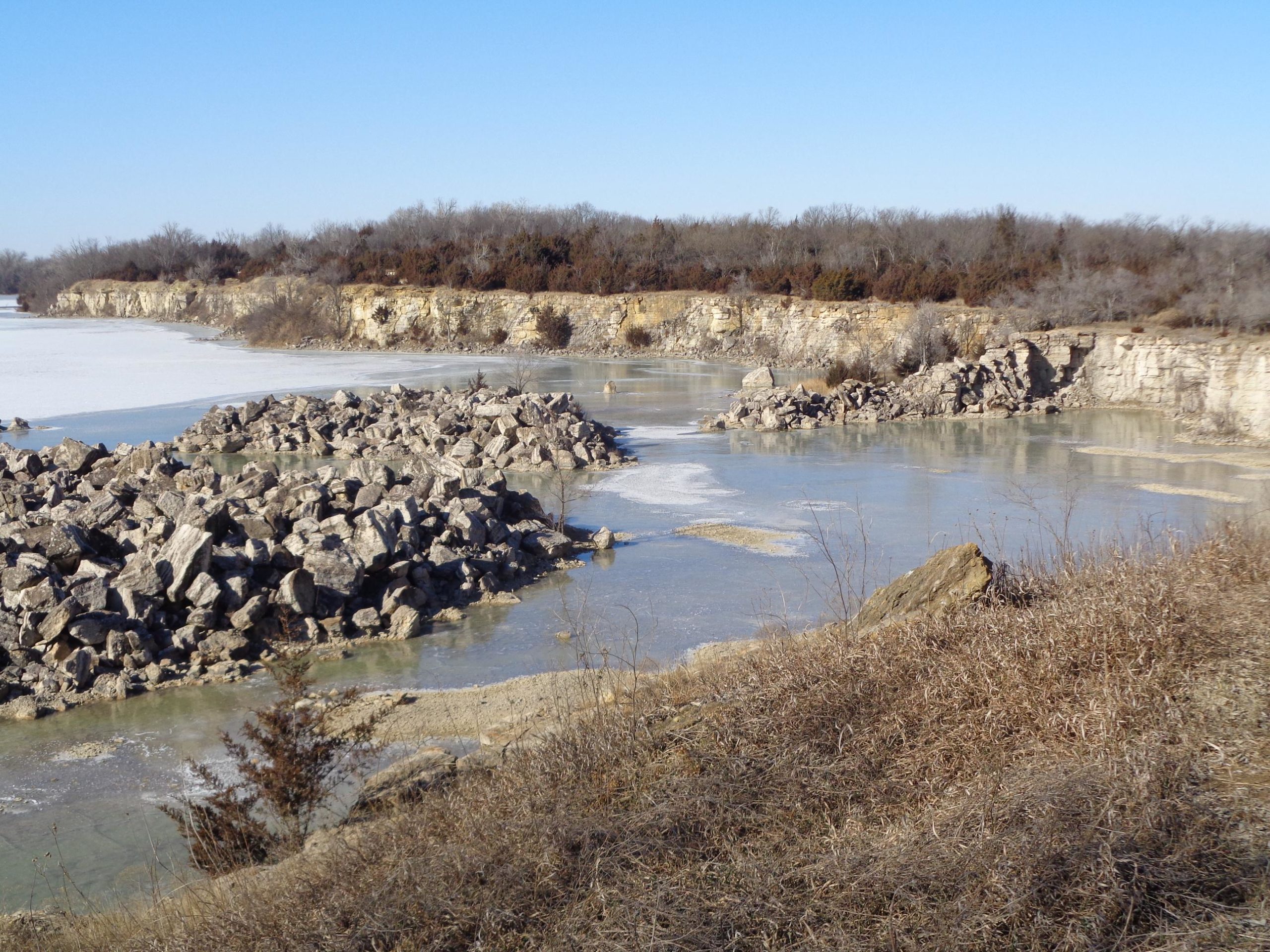 A scenic view of a frozen lake surrounded by rocky shorelines and barren trees under a clear blue sky. Large boulders can be seen gathered along the water's edge, with patches of ice on the lake’s surface. The landscape features a mix of brown grasses and rocky cliffs in the background. Lehigh Portland Trails mountain bike trail.
