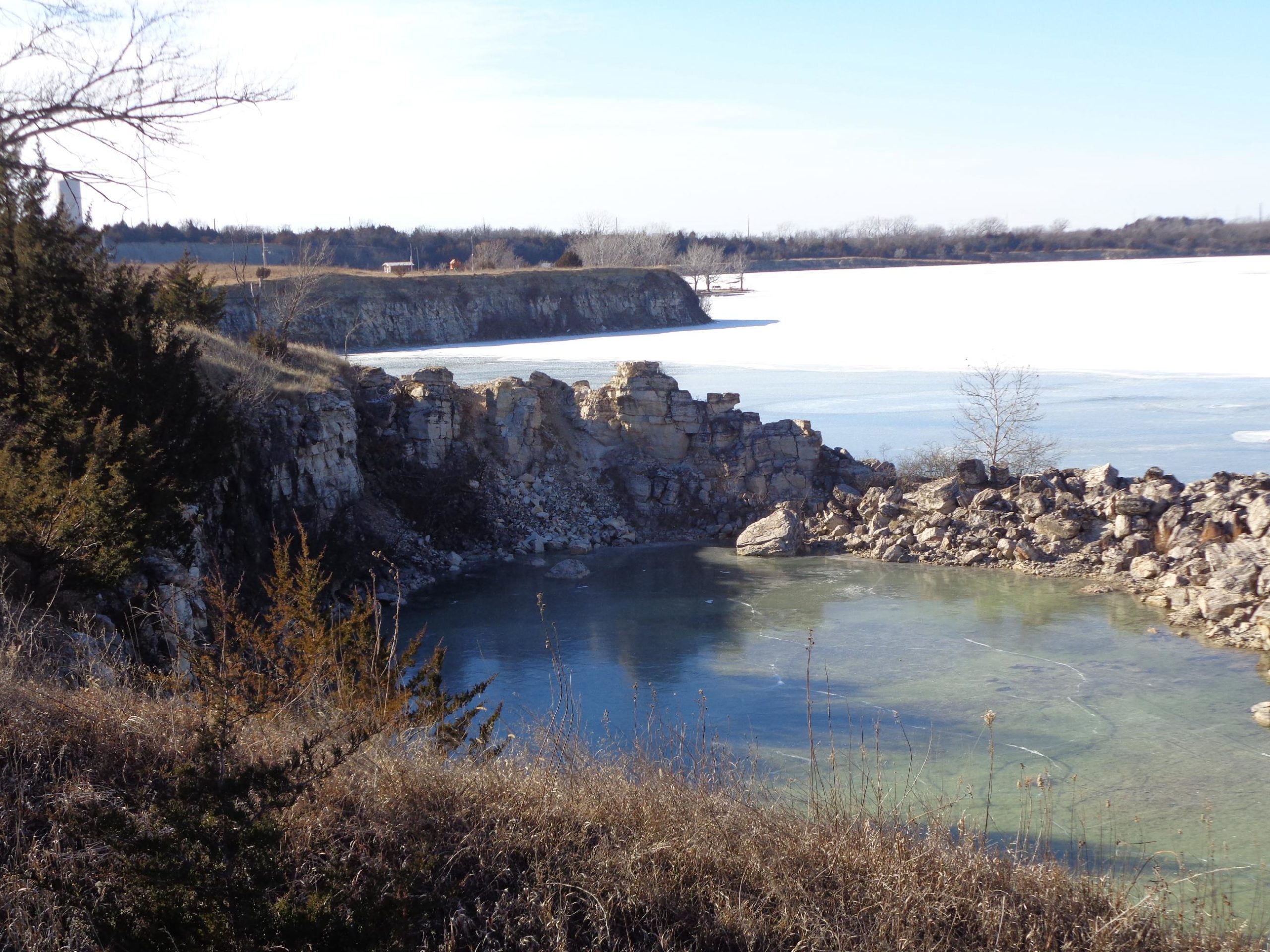A scenic view of rocky cliffs beside a calm body of water, with a partially frozen lake in the background under a clear blue sky. The foreground features dry grass and scattered shrubs, while the cliffs are made up of layered stone formations. Lehigh Portland Trails mountain bike trail.