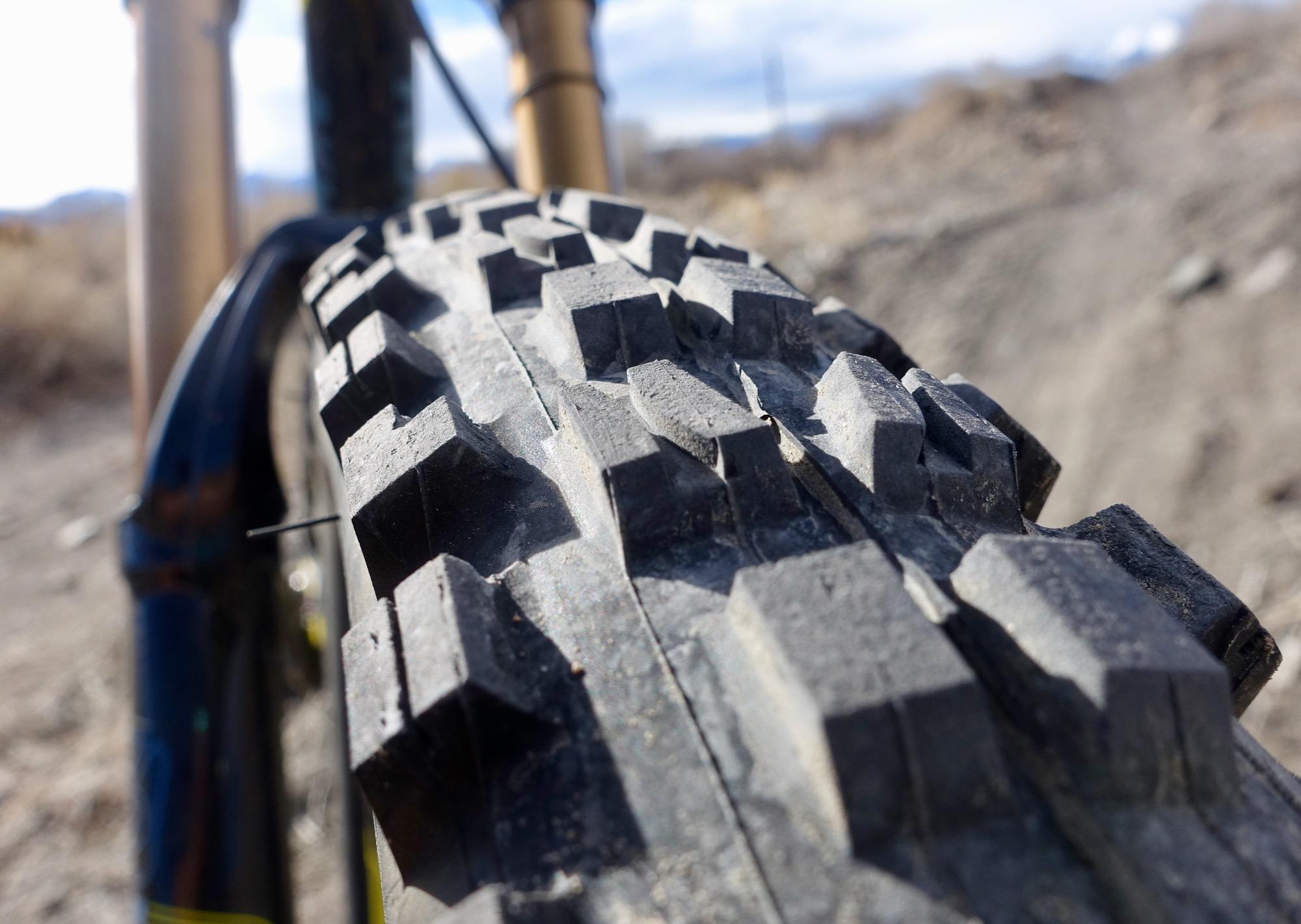 Maxxis Minion DHF: Close-up view of a textured mountain bike tire with deep treads, set against a blurred background of dirt and sparse vegetation, showcasing the tire's grip and rugged design.