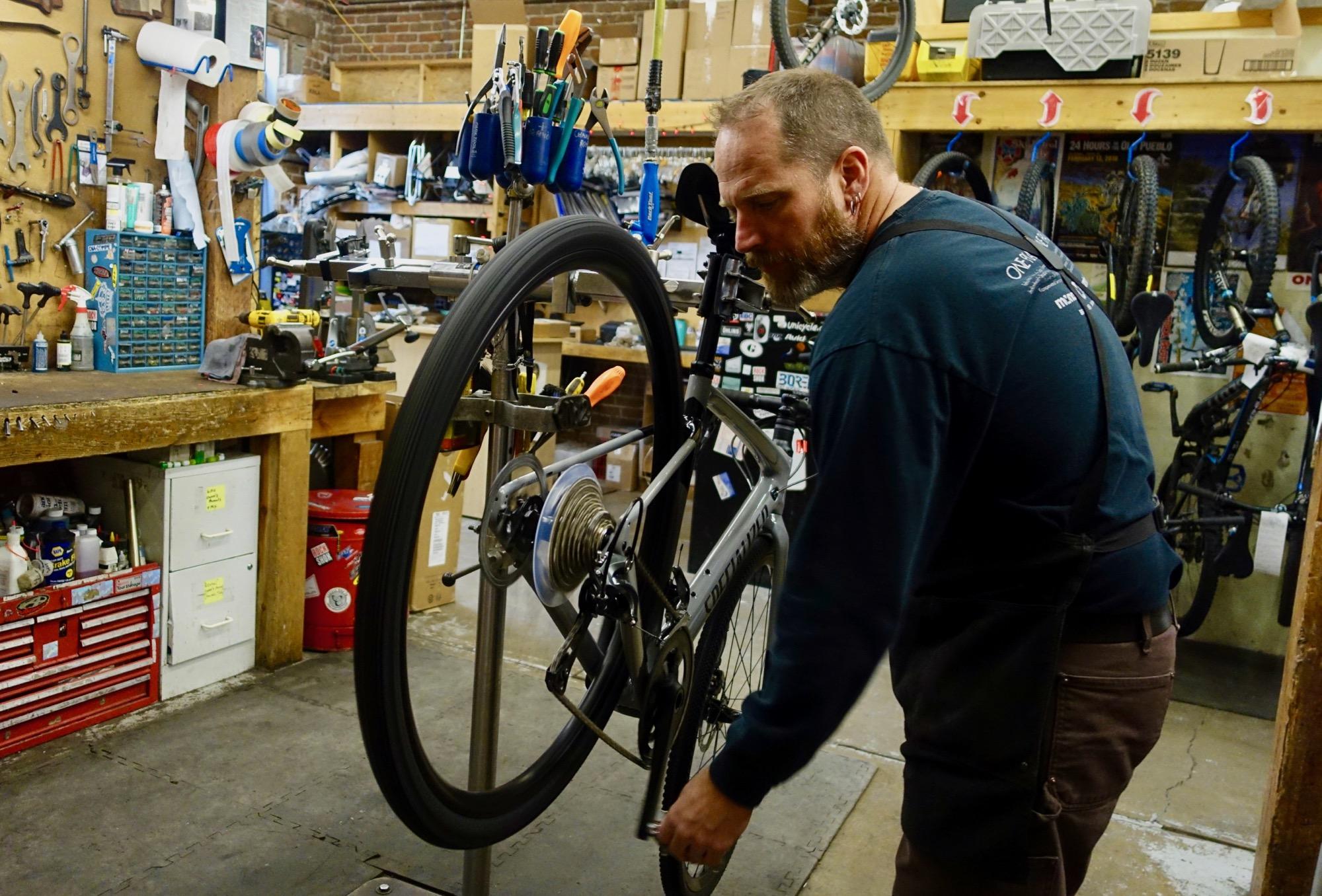 A bicycle mechanic working on a bike in a cluttered workshop filled with tools and bike parts. The mechanic is focused on adjusting the bike's drivetrain while surrounded by various tools hanging on the wall and additional bikes in the background.