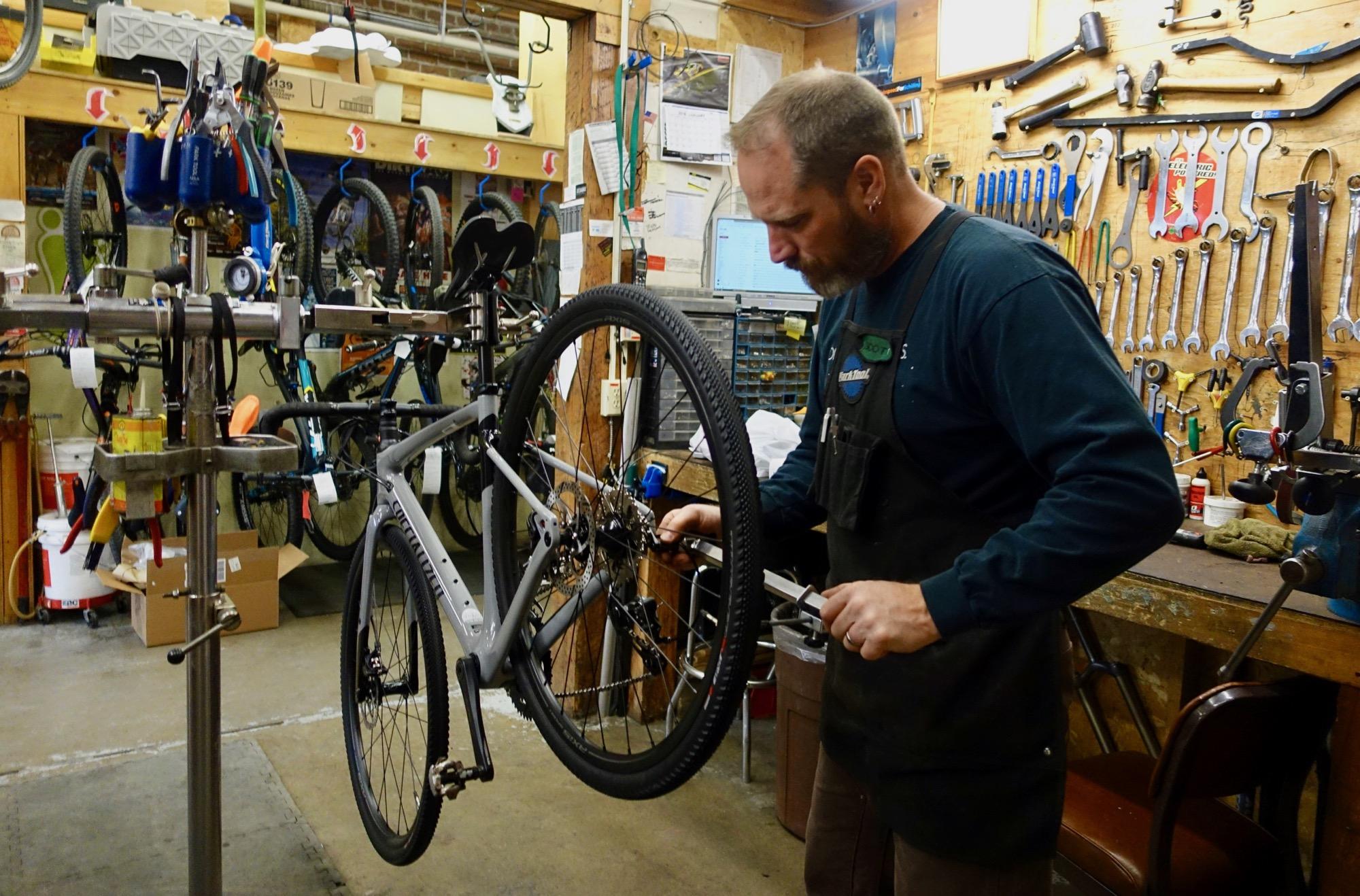 A mechanic working on a bicycle in a workshop filled with tools and parts. The bike is mounted on a repair stand, and the mechanic is focused on adjusting the rear wheel using a wrench. The background features various tools hanging on the wall and additional bikes stored on racks.