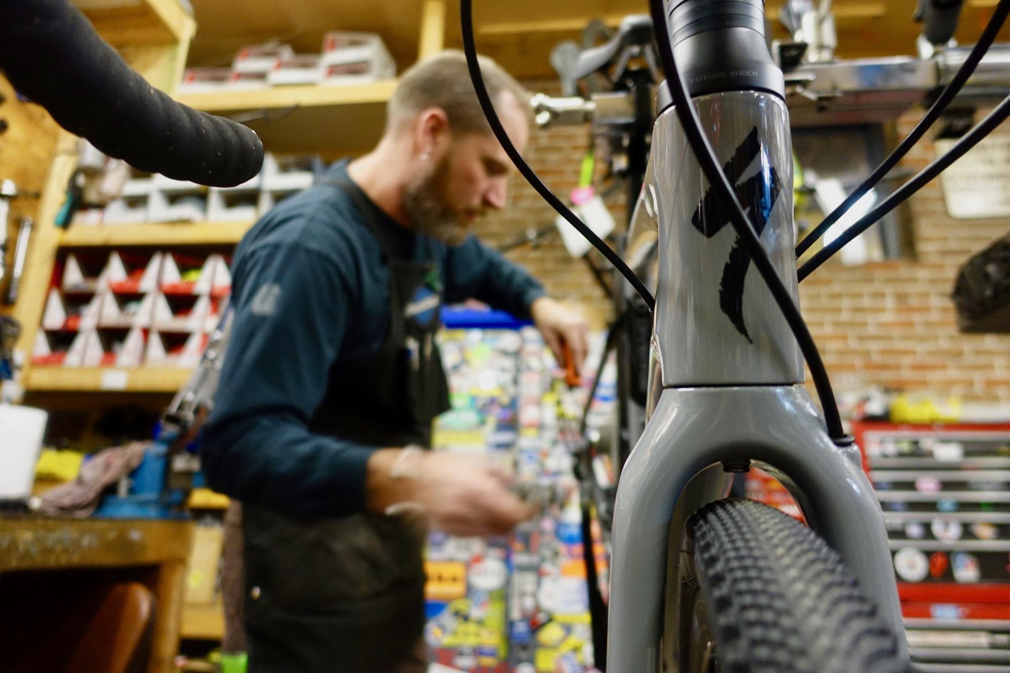 A bicycle mechanic working in a workshop, focused on adjusting a bike frame. The foreground shows a close-up of the bike's frame and handlebars, while the background features shelves filled with bike parts and tools. The mechanic is visible with a beard and is wearing an apron, engaged in his task.