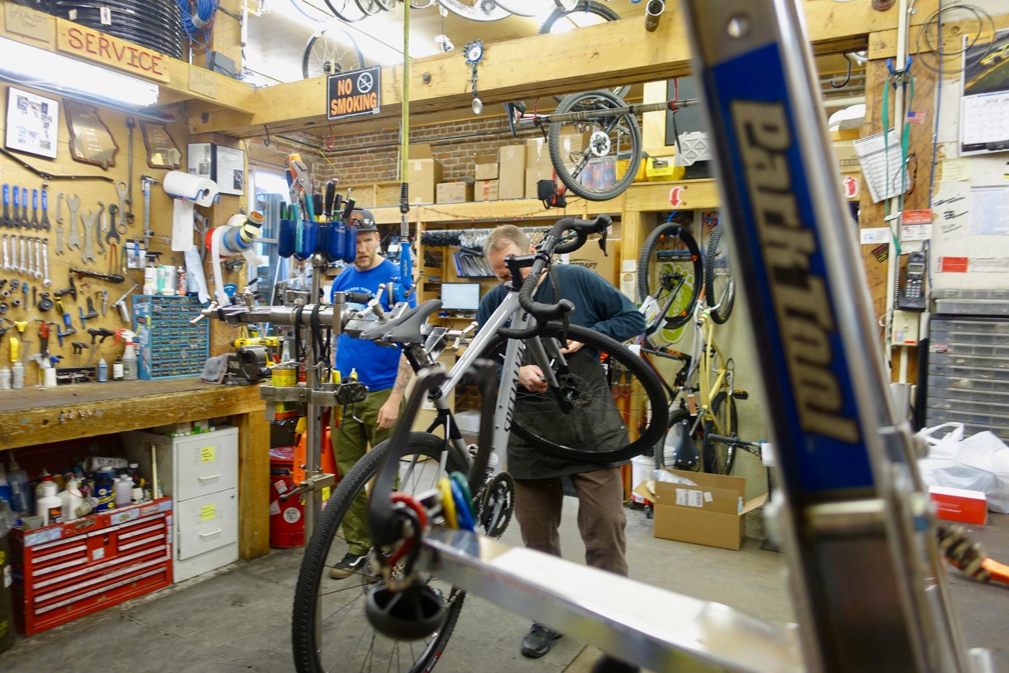 A bike repair shop featuring two mechanics working on bicycles. One mechanic is adjusting a bike in the foreground while the other is observing. The walls are lined with various bike tools, and there are bicycles hanging from the ceiling. A sign that reads "NO SMOKING" is visible in the background.