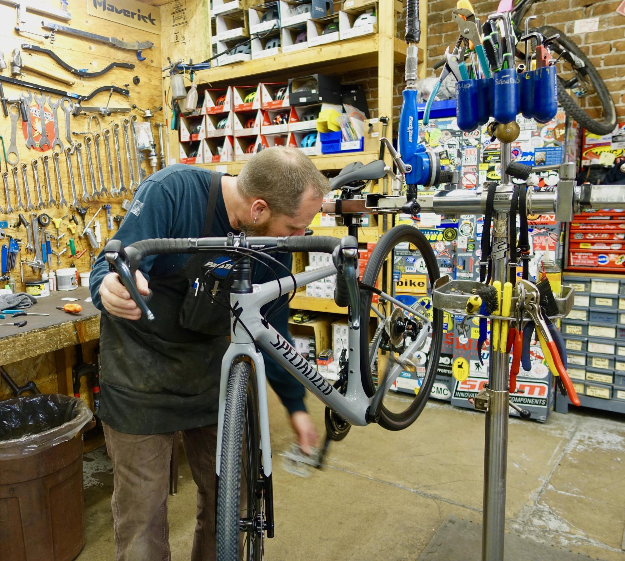 A mechanic is working on a bicycle in a workshop. The bike is mounted on a repair stand, and the mechanic is using a cloth to clean the frame. Various tools, including wrenches and screwdrivers, are neatly organized on the wall and on the workbench, highlighting the workshop's gear and bike repair environment.