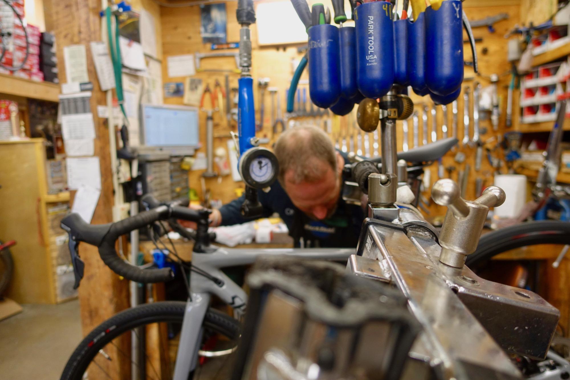 A bicycle mechanic working in a workshop, focused on repairing a bike. Tools and equipment are organized on the walls, while a workspace with a bike frame is visible in the foreground.