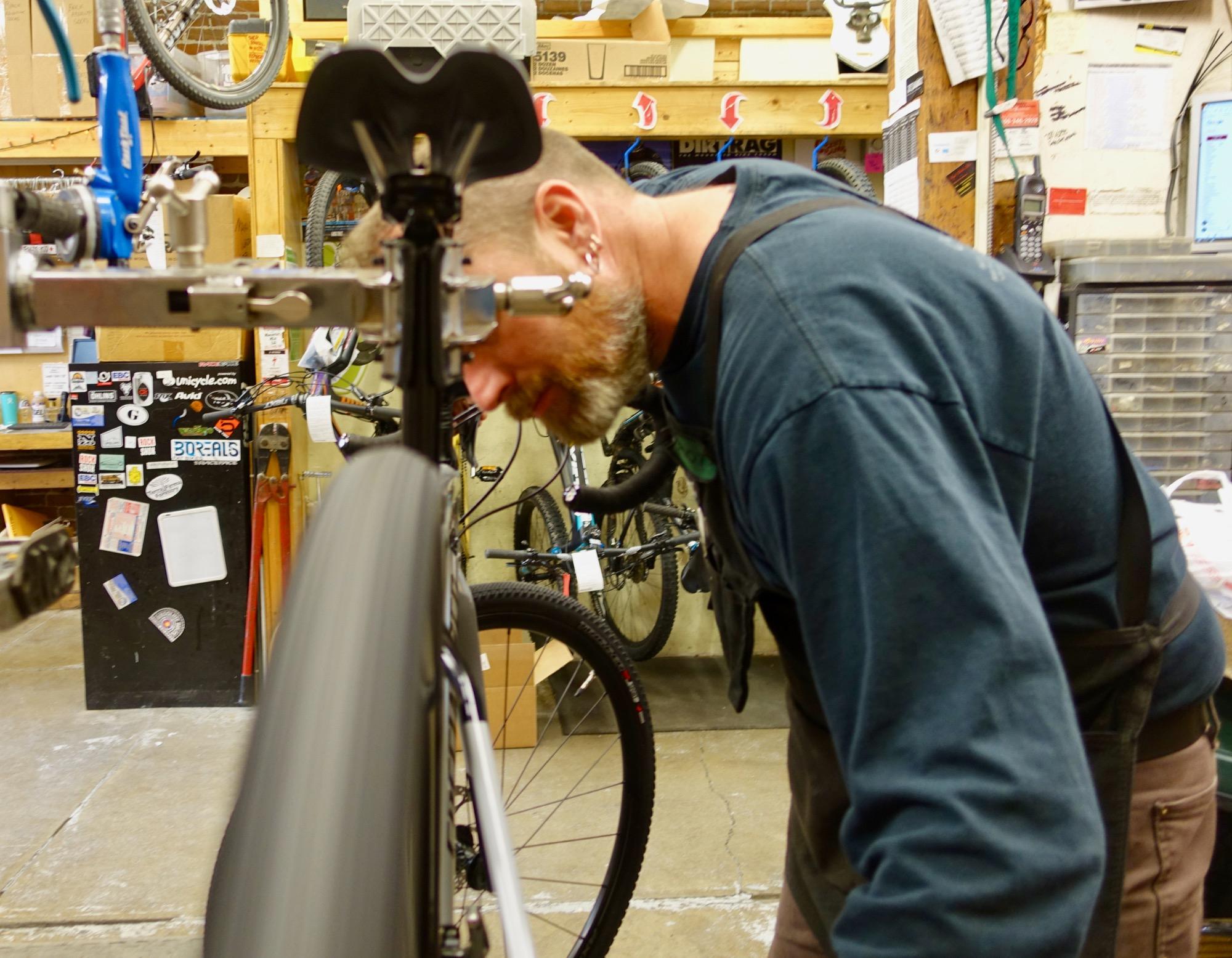 A mechanic working on a bicycle in a workshop, leaning close to adjust a component while a bike tire spins in front of him. The background features various bikes and tools, with a cluttered workbench displaying stickers and equipment.