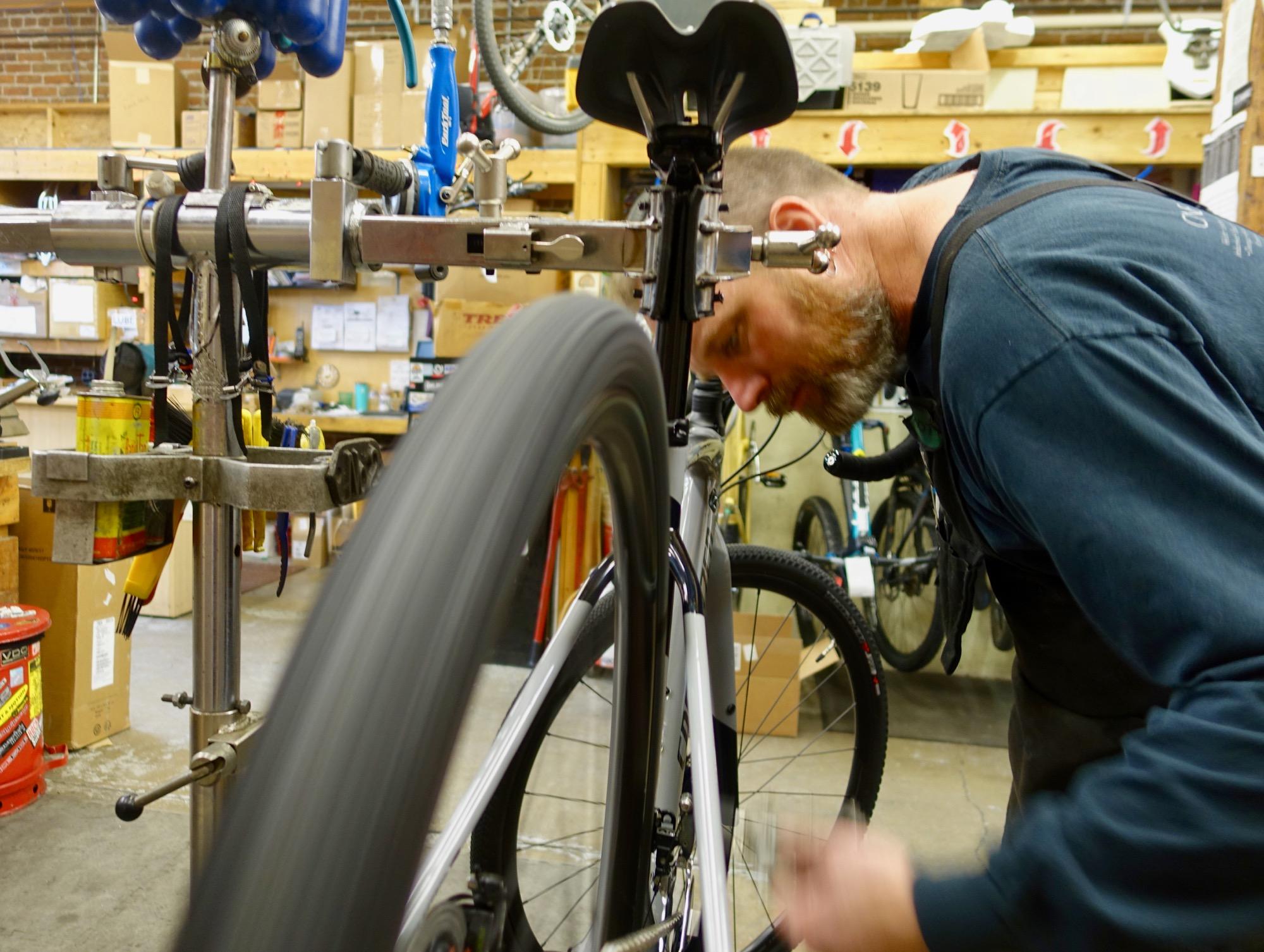A mechanic working on a bicycle in a workshop, focusing on the rear tire while using tools. The background includes various bikes and equipment, highlighting a busy bike repair environment.