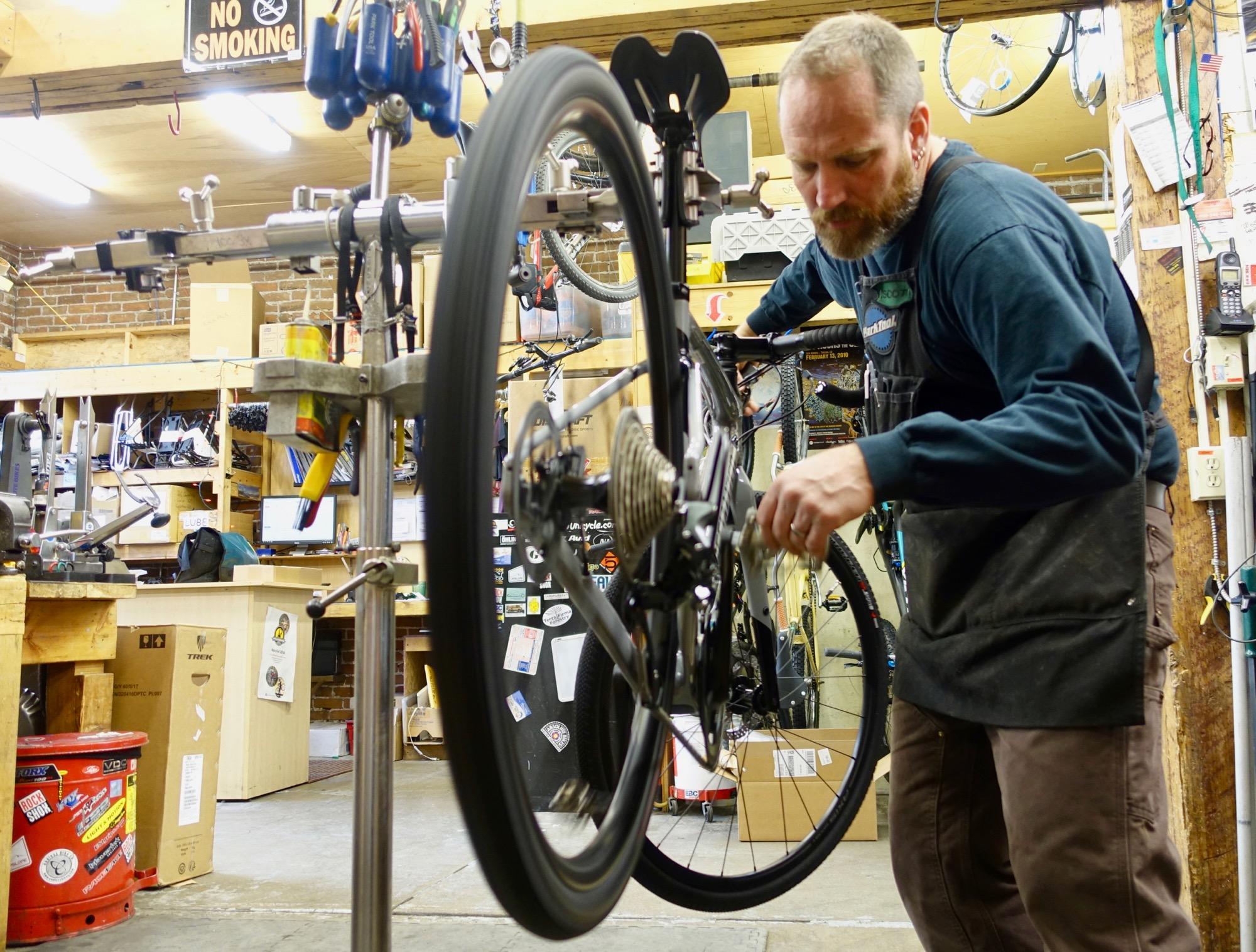 A bicycle mechanic works on a bike in a workshop, adjusting the gears while the bike is mounted on a repair stand. The background features shelves filled with bicycle parts and tools, creating a busy and professional atmosphere.