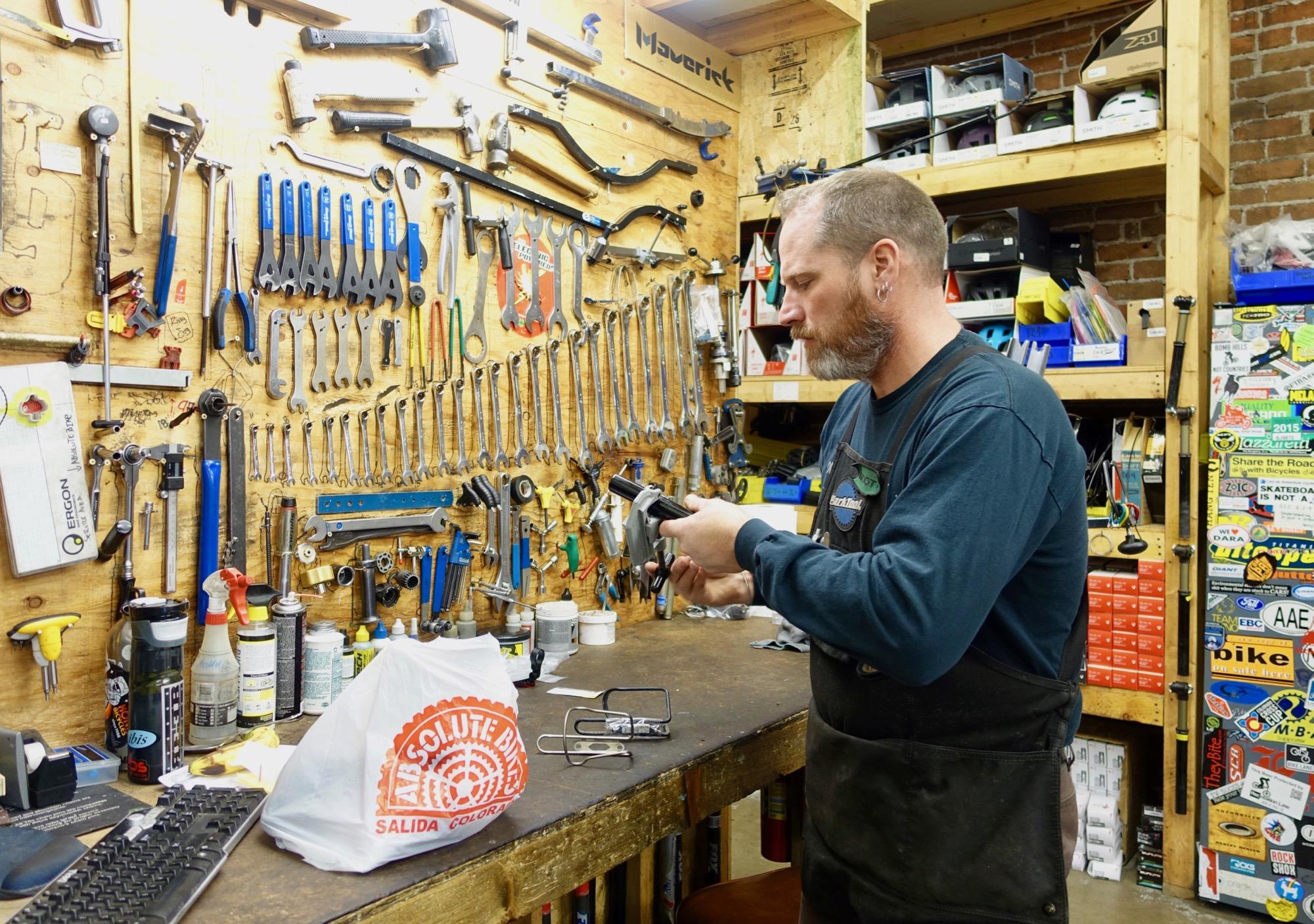 A workshop scene featuring a person examining a tool while standing at a workbench cluttered with various hand tools and supplies. A wall-mounted display showcases an array of wrenches and other tools. In the foreground, there is a bag from a local bike shop and a keyboard on the workbench. The space is organized yet filled with items indicative of a busy repair or maintenance environment.