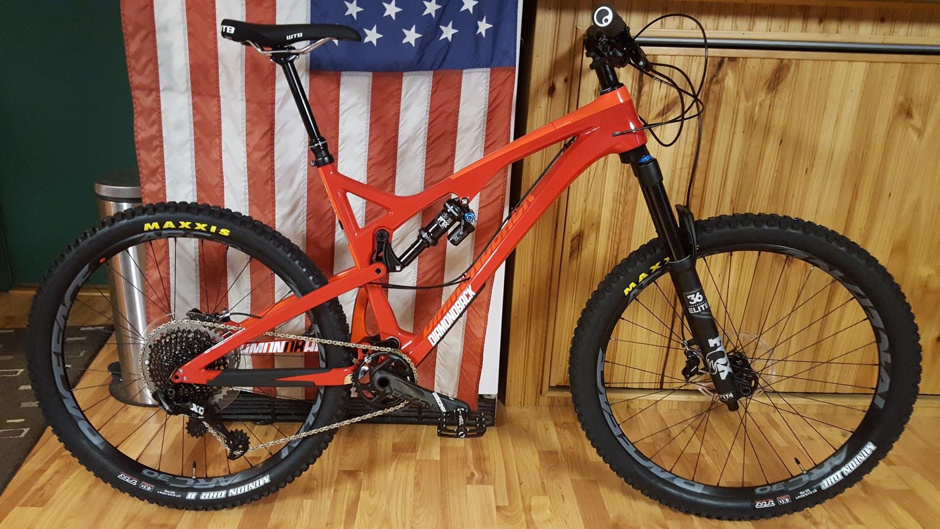 Diamondback Release 5c: A vibrant red mountain bike with a lightweight frame and prominent suspension system stands on a wooden floor, positioned in front of a backdrop featuring an American flag. The bike showcases oversized tires, one with the brand "Maxxis" prominently displayed, and a modern design suitable for off-road cycling.