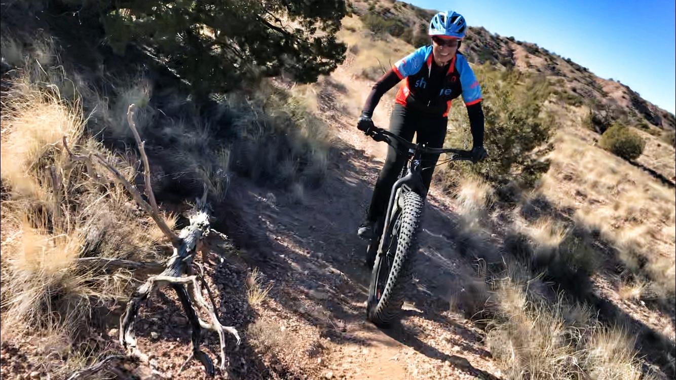 A person riding a mountain bike through a rugged trail, surrounded by dry grass and sparse vegetation. The cyclist is wearing a blue and black helmet and a colorful jersey, smiling as they navigate the terrain. A sunlit landscape features hills in the background. Mariposa Fat Bike Trails mountain bike trail.