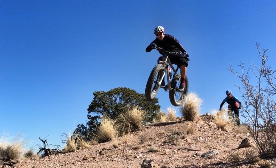 A person wearing a helmet and cycling gear jumps off a dirt incline while riding a mountain bike. In the background, another cyclist is visible riding up the slope. The scene is set against a clear blue sky with sparse vegetation and rocky terrain. Mariposa Fat Bike Trails mountain bike trail.
