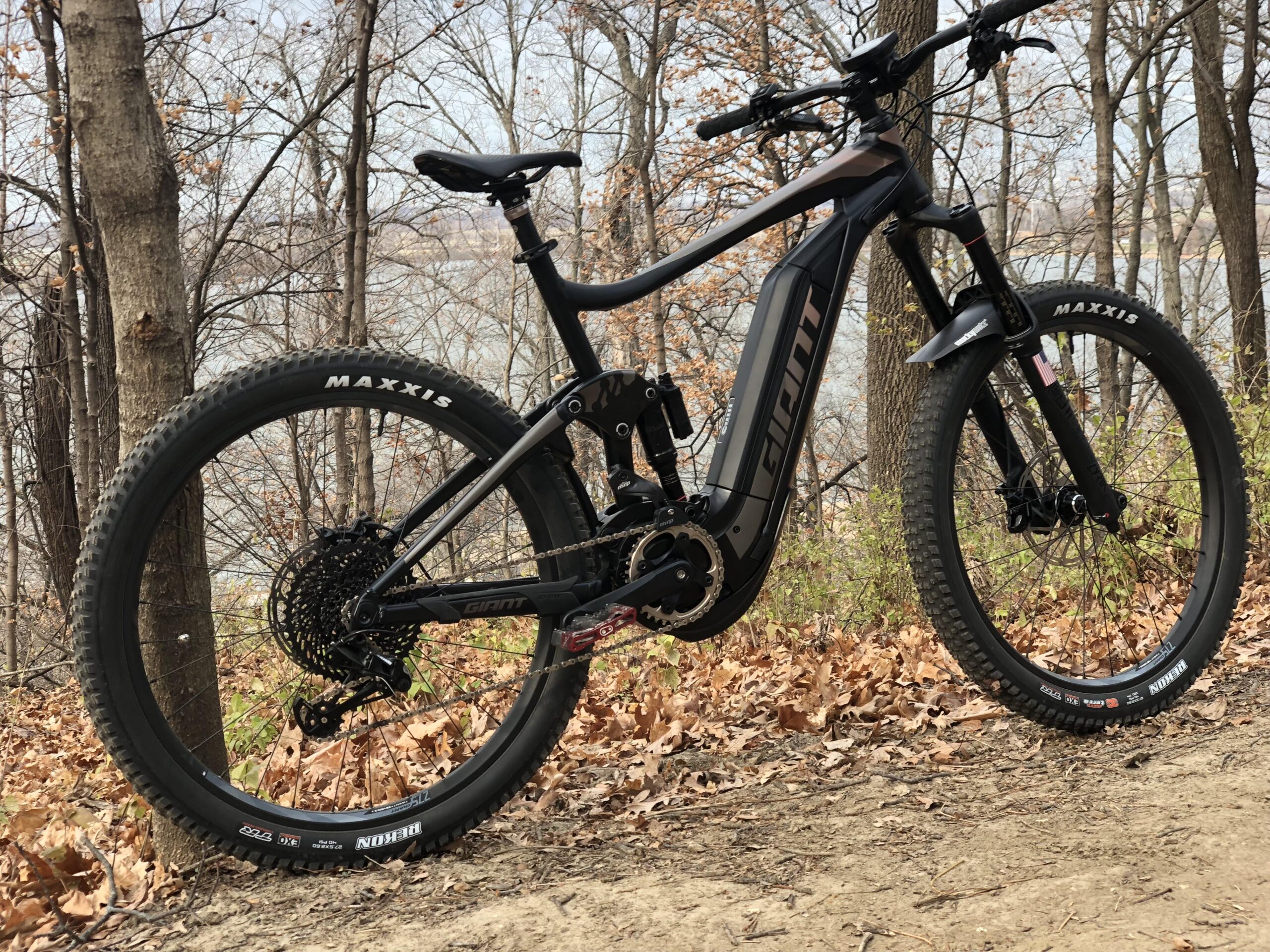 Giant FULL-E  0 SX PRO: A black electric mountain bike positioned on a dirt path surrounded by trees and dry foliage. The bike features thick tires with "Maxxis" branding, a prominent battery pack, and the brand name "Giant" displayed prominently on the frame. In the background, a body of water is visible through the trees.
