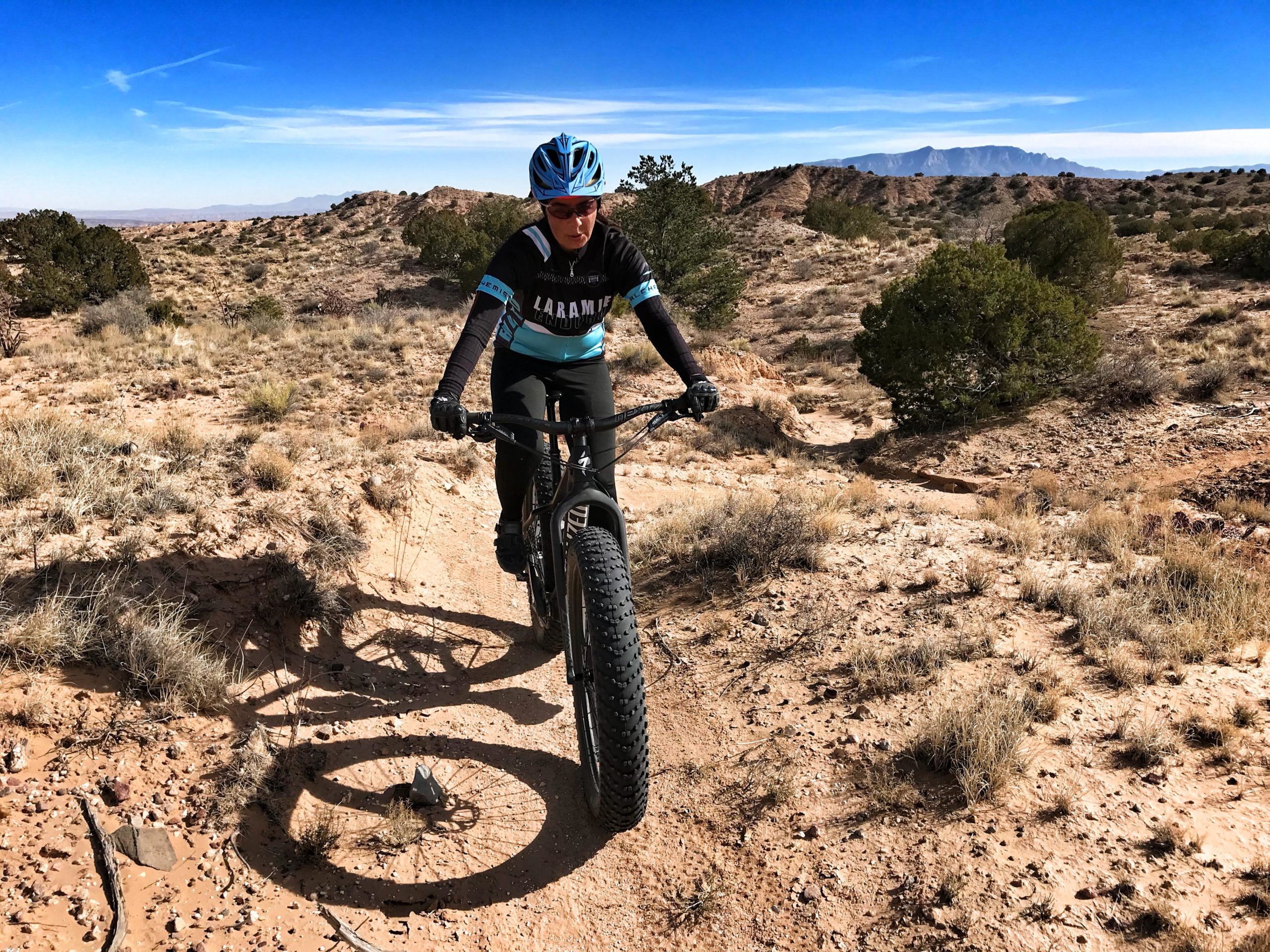 A person riding a fat bike along a sandy trail in a desert landscape, surrounded by sparse vegetation and rocky hills under a clear blue sky. The rider wears a blue helmet and a cycling jersey, focused on navigating the terrain. Mariposa Fat Bike Trails mountain bike trail.